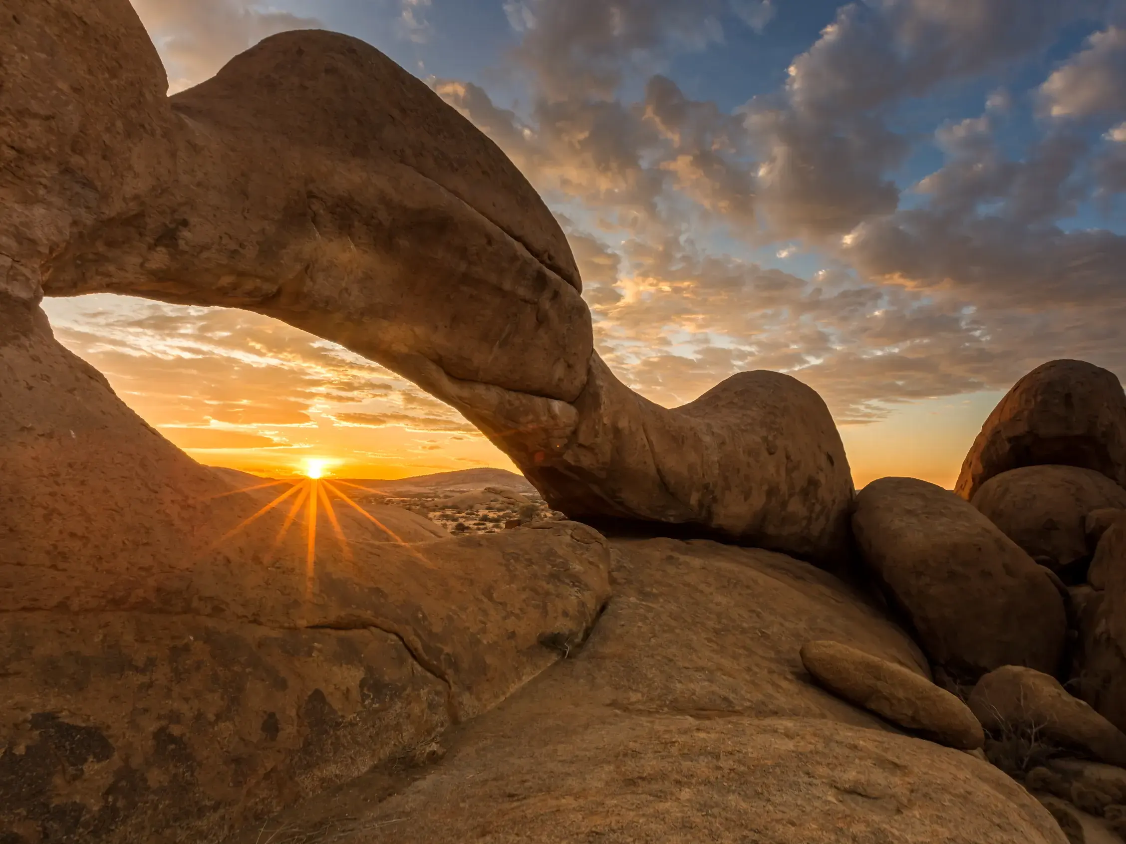 Sonnenuntergang mit Sonnenstern durch den markanten Felsbogen der Spitzkoppe in der weiten Wüstenlandschaft Namibias.
