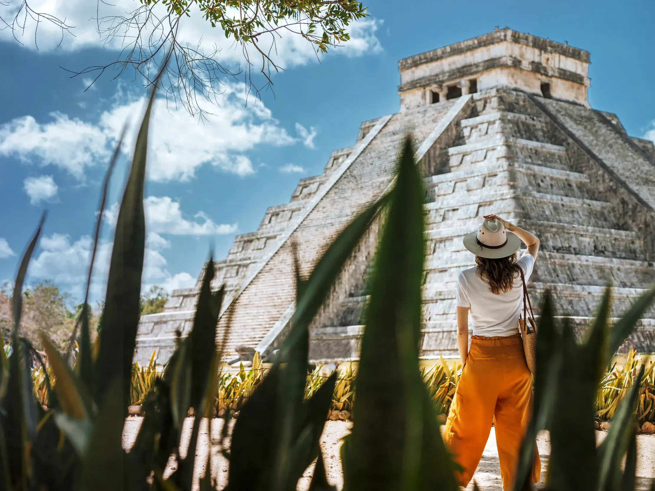 Eine Reisende mit Hut blickt hinter grünen Agaven empor zur mächtigen Maya-Pyramide von Chichén Itzá auf der Yukatan-Halbinsel.