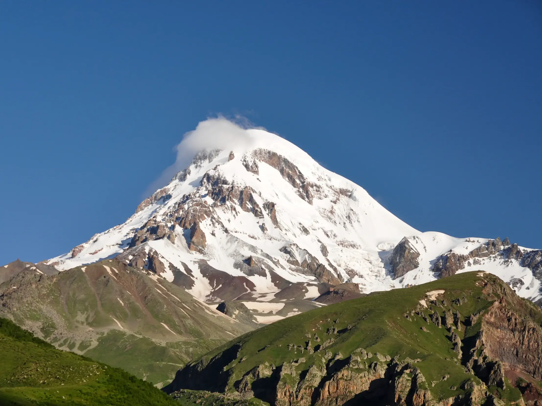 Majestätischer Blick auf den schneebedeckten Gipfel des Kasbek in Georgien, im Vordergrund die grünen Hügel des Kaukasus.