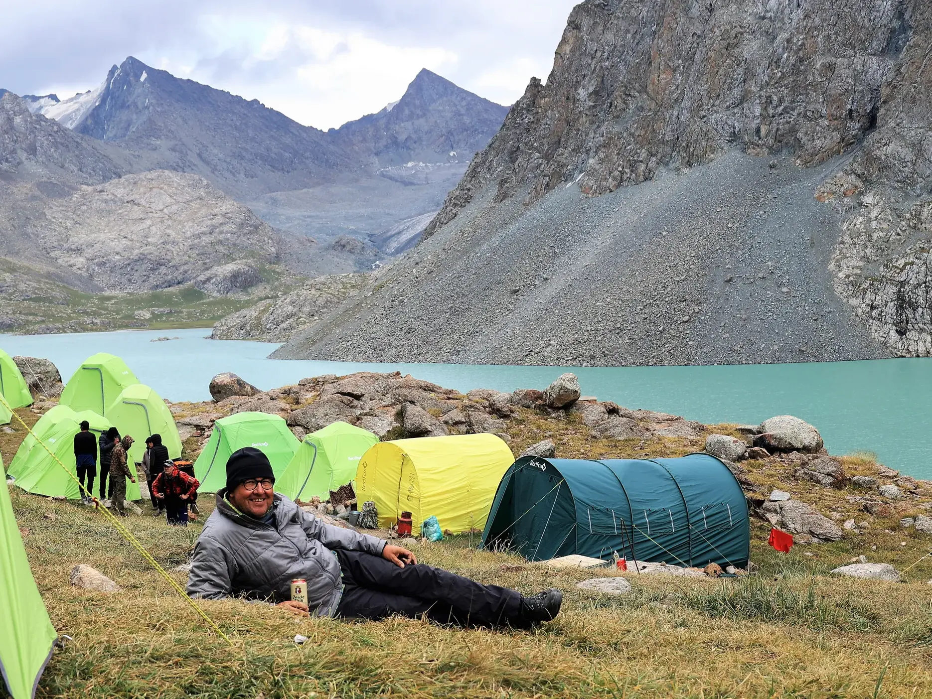 Ein Wanderer entspannt im Zeltlager am Ufer des türkisen Sees Alaköl vor der Bergkulisse des Terskej-Alatau-Gebirges.