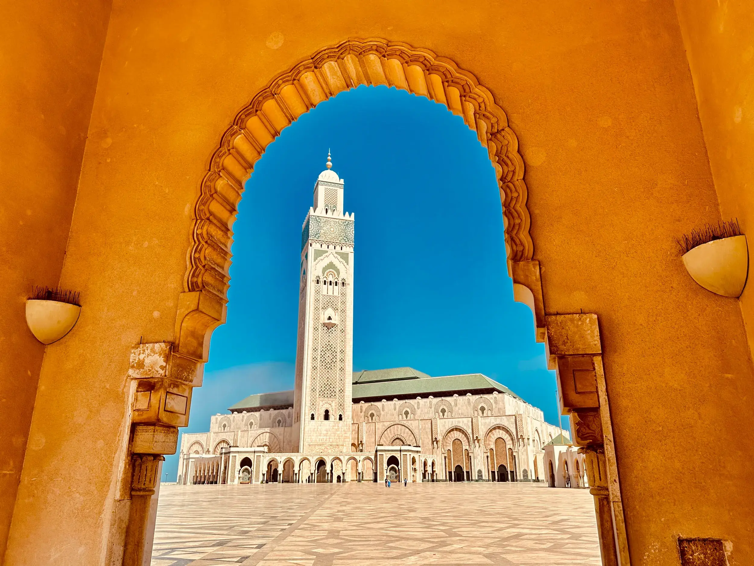 Prachtvoller Blick durch einen ockerfarbenen Torbogen auf das Minarett der großen Hassan-II.-Moschee in Casablanca vor blauem Himmel.