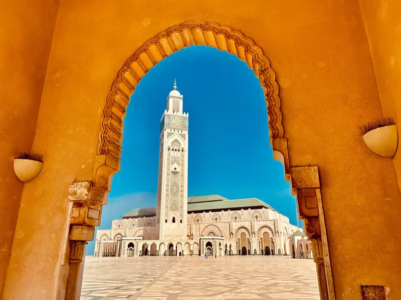 Prachtvoller Blick durch einen ockerfarbenen Torbogen auf das Minarett der großen Hassan-II.-Moschee in Casablanca vor blauem Himmel.