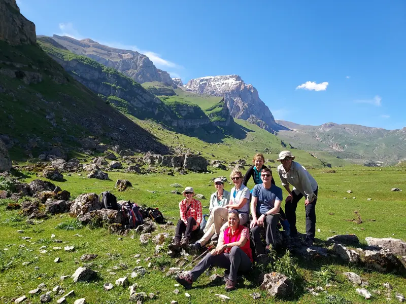 Eine siebenköpfige Wandergruppe pausiert lächelnd auf einer Wiese vor dem schneebedeckten Berg Shahdag im Großen Kaukasus.