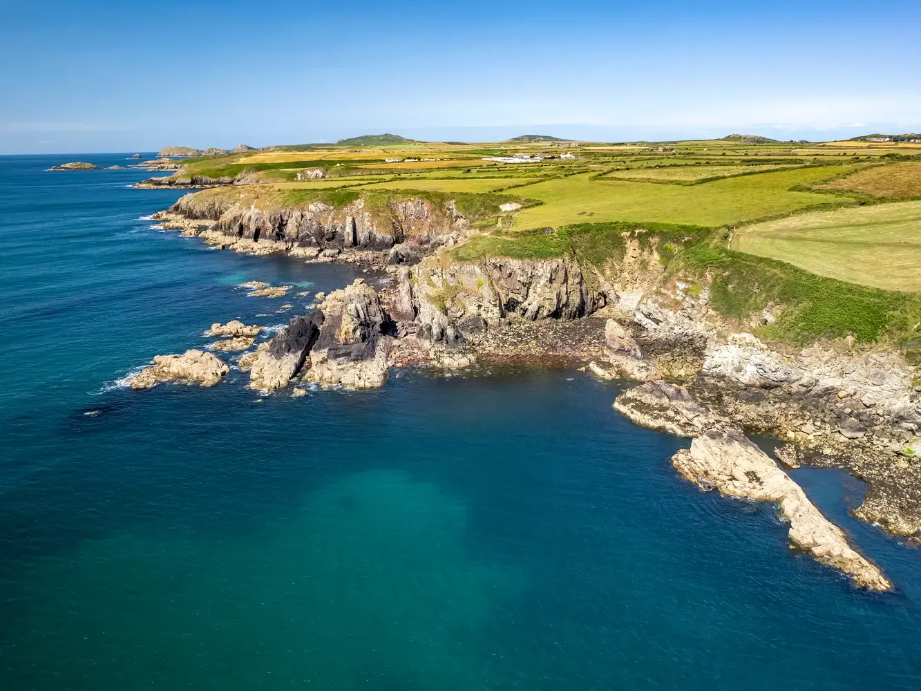 Sonnige Luftaufnahme der zerklüfteten Felsenküste am Pembrokeshire Coast Path mit grünen Wiesen und türkisblauem Meer in Wales.