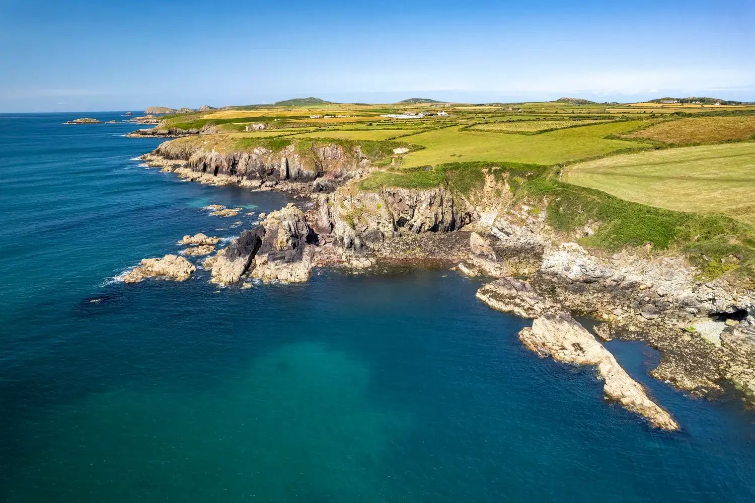 Sonnige Luftaufnahme der zerklüfteten Felsenküste am Pembrokeshire Coast Path mit grünen Wiesen und türkisblauem Meer in Wales.