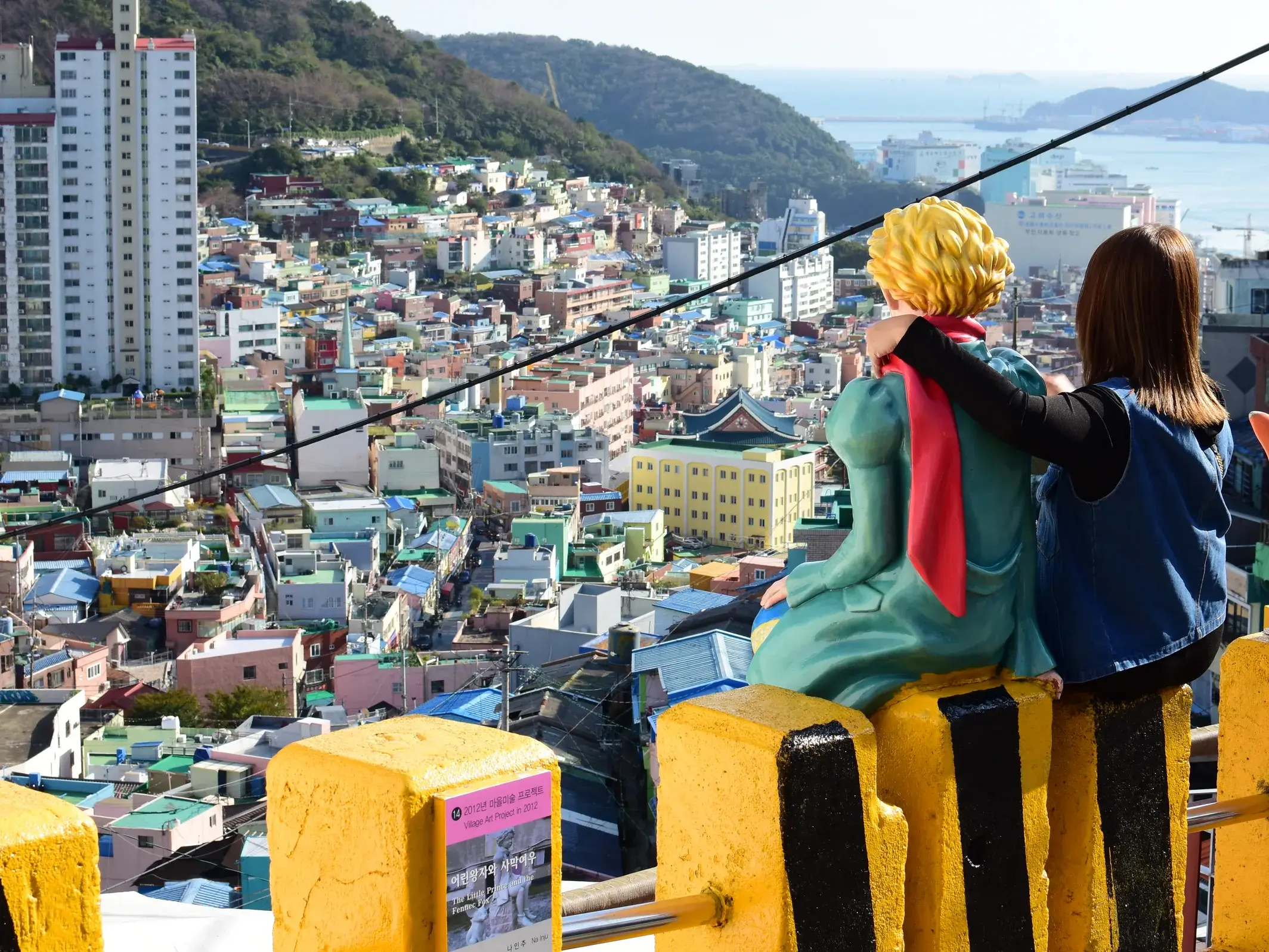 Junge Frau legt den Arm um die Statue des Kleinen Prinzen und blickt auf das bunte Gamcheon Culture Village in Busan.
