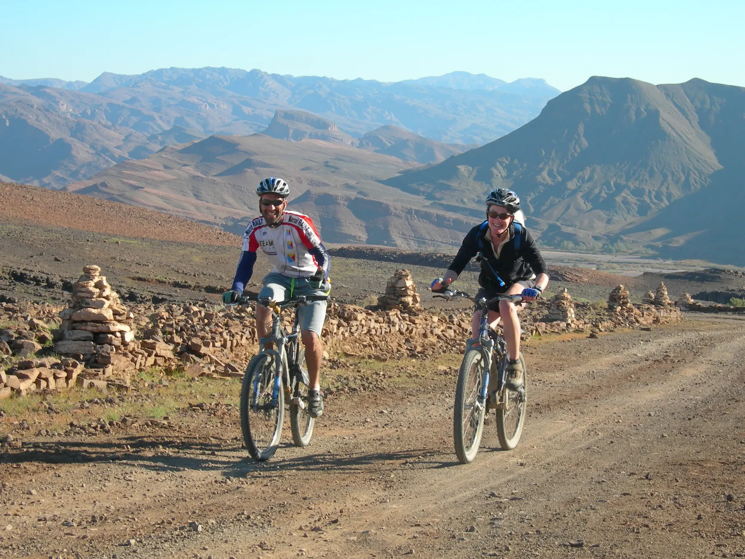 Zwei lächelnde Mountainbiker auf einer Schotterpiste während einer Gruppenreise durch das Jebel Sarhro Gebirge in Marokko.