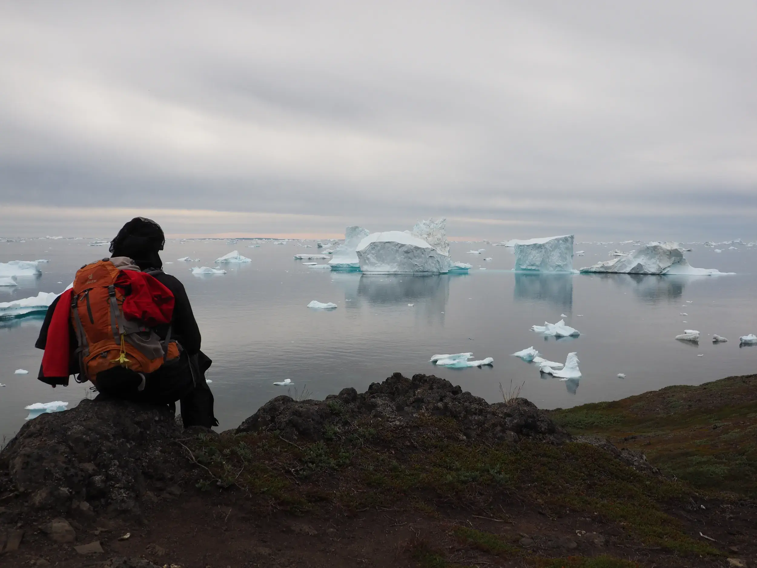 Wanderer mit Trekkingrucksack macht Pause auf Felsen mit Blick auf die gigantischen Eisberge des Ilulissat-Eisfjords, Grönland.