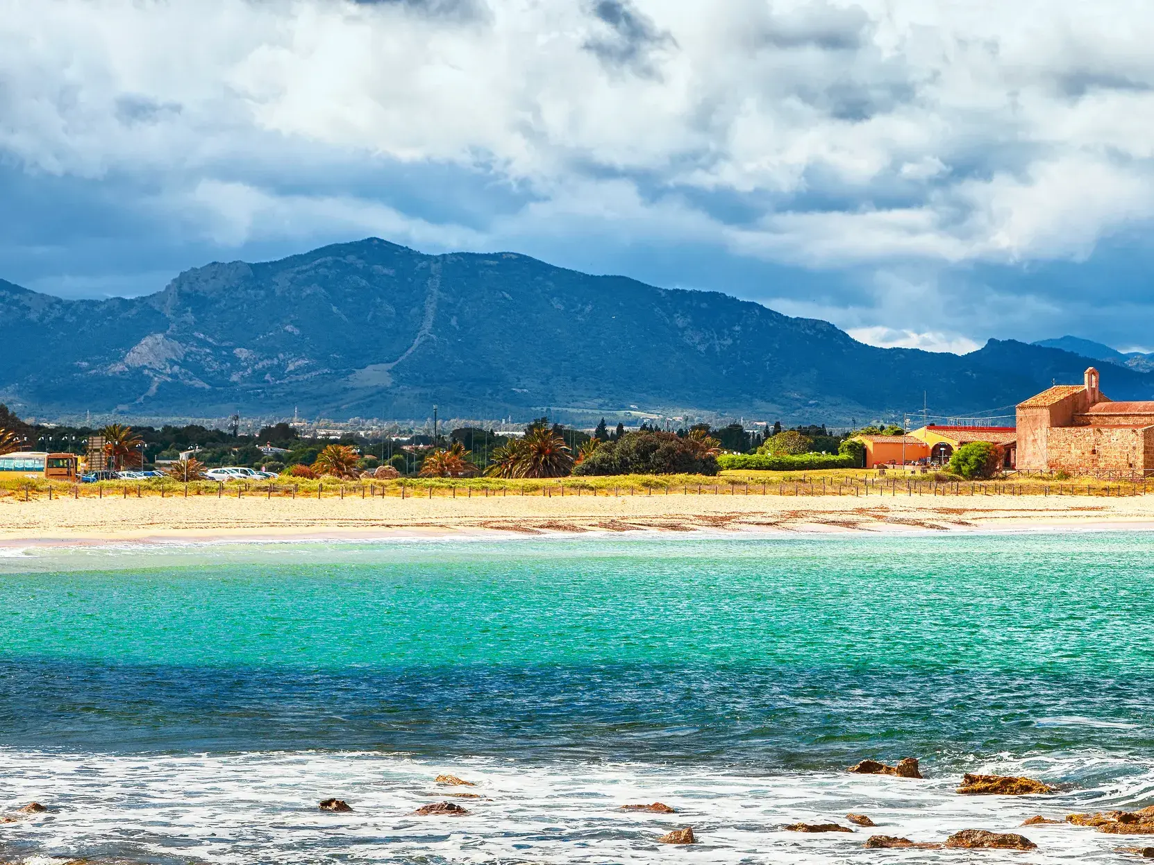 Historische Kirche Sant'Efisio am Strand von Nora auf Sardinien, davor türkises Meer, im Hintergrund eine Bergkulisse.