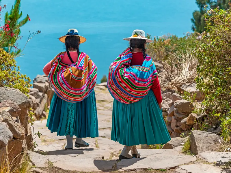 Zwei Frauen in traditioneller Anden-Tracht mit bunten Tragetüchern gehen einen Steinpfad auf der Isla del Sol am Titicacasee hinab.