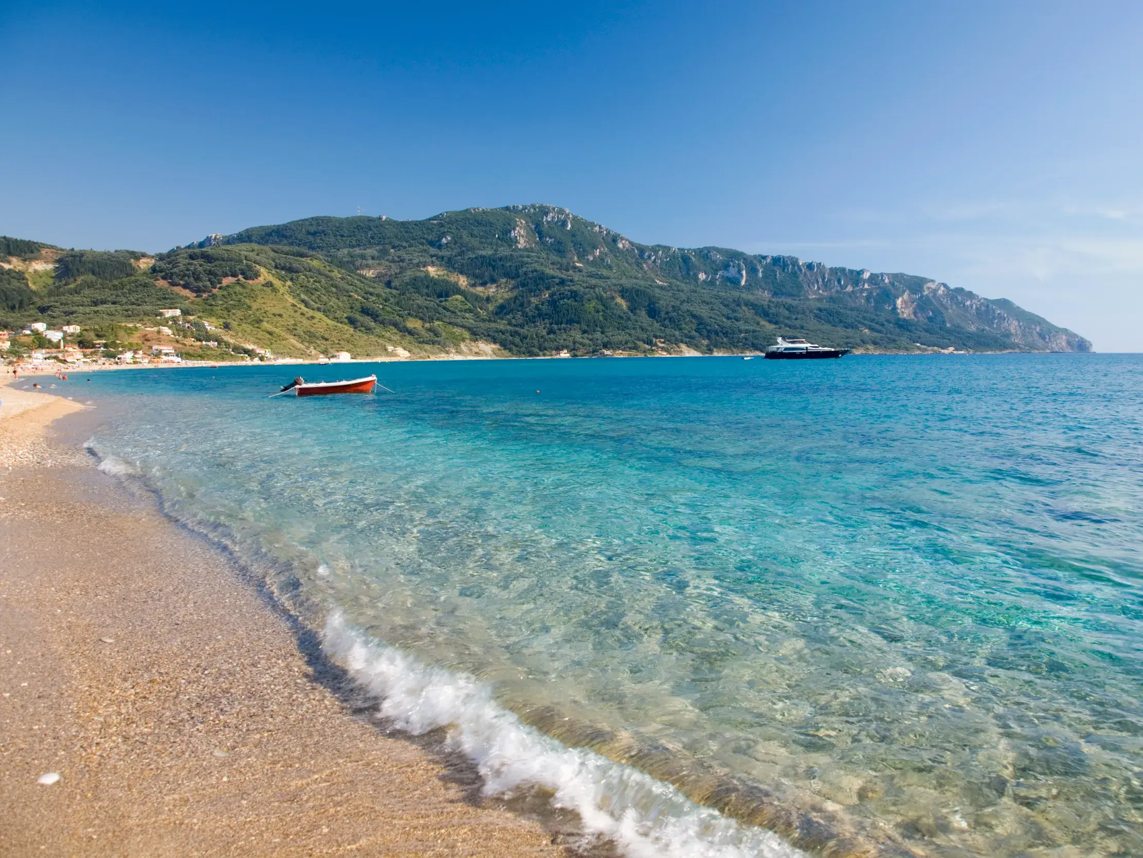 Glasklares, türkises Wasser umspült den Sandstrand einer Bucht auf Korfu, im Hintergrund die grüne, hügelige Küste.