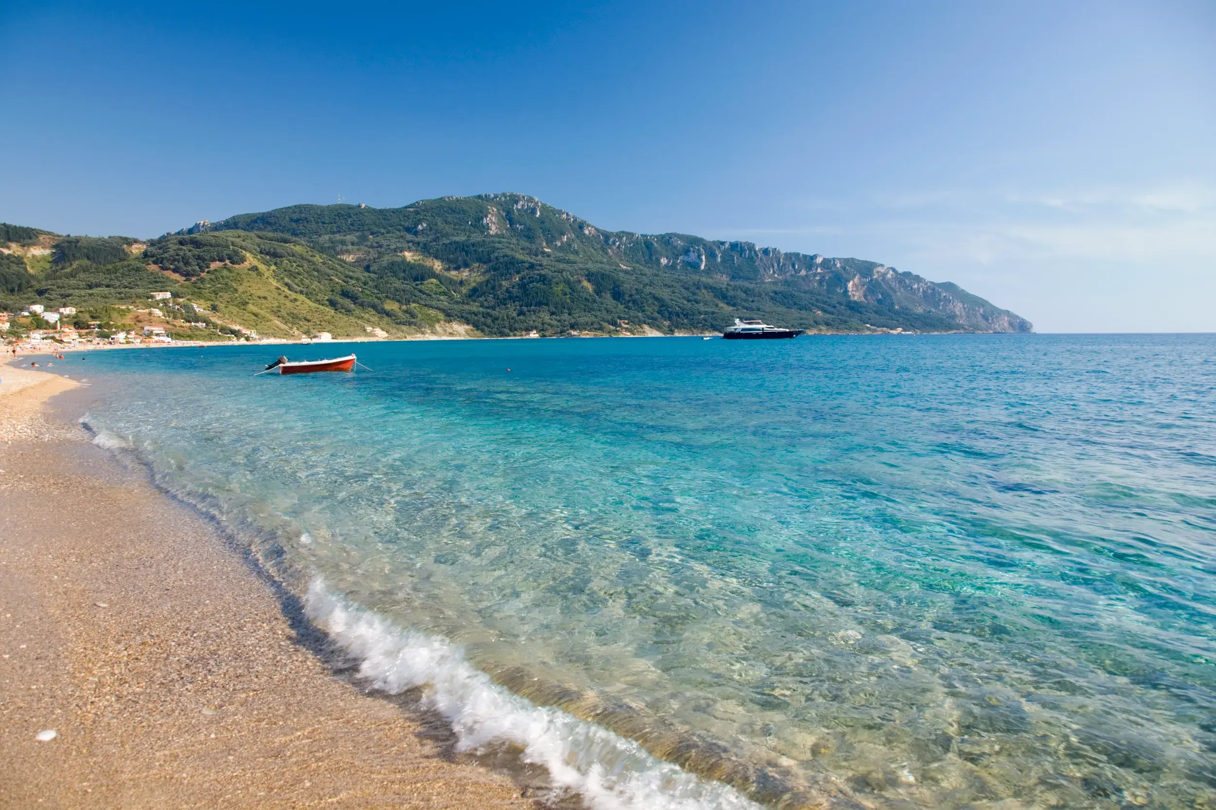 Glasklares, türkises Wasser umspült den Sandstrand einer Bucht auf Korfu, im Hintergrund die grüne, hügelige Küste.
