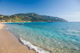 Glasklares, türkises Wasser umspült den Sandstrand einer Bucht auf Korfu, im Hintergrund die grüne, hügelige Küste.