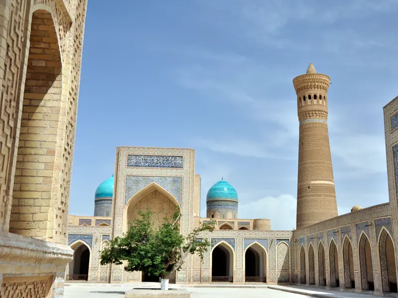 Sonniger Innenhof des Poi Kalyan Komplexes in Buchara mit dem Kalyan-Minarett und blauen Kuppeln der Mir-i-Arab-Medrese.