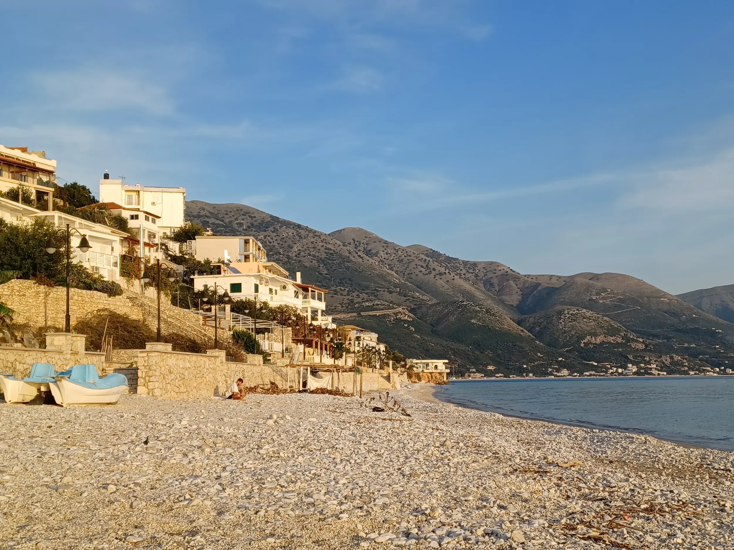 Weitläufiger Kieselstrand in Himare an der Albanischen Riviera mit weißen Häusern am Hang vor der Bergkulisse am Abend.