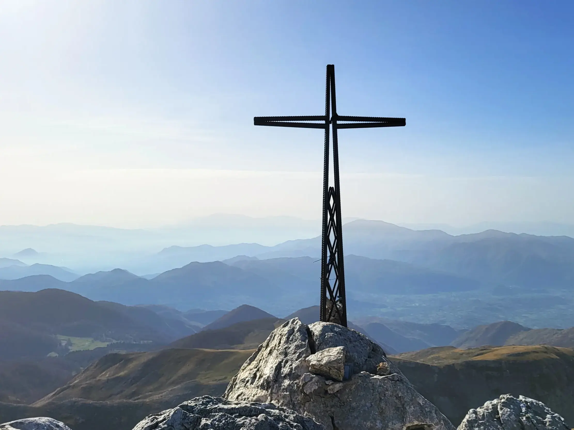 Metallenes Gipfelkreuz auf dem felsigen Gipfel des Monte Meta mit Panoramablick über die blauen Bergketten des Apennin.