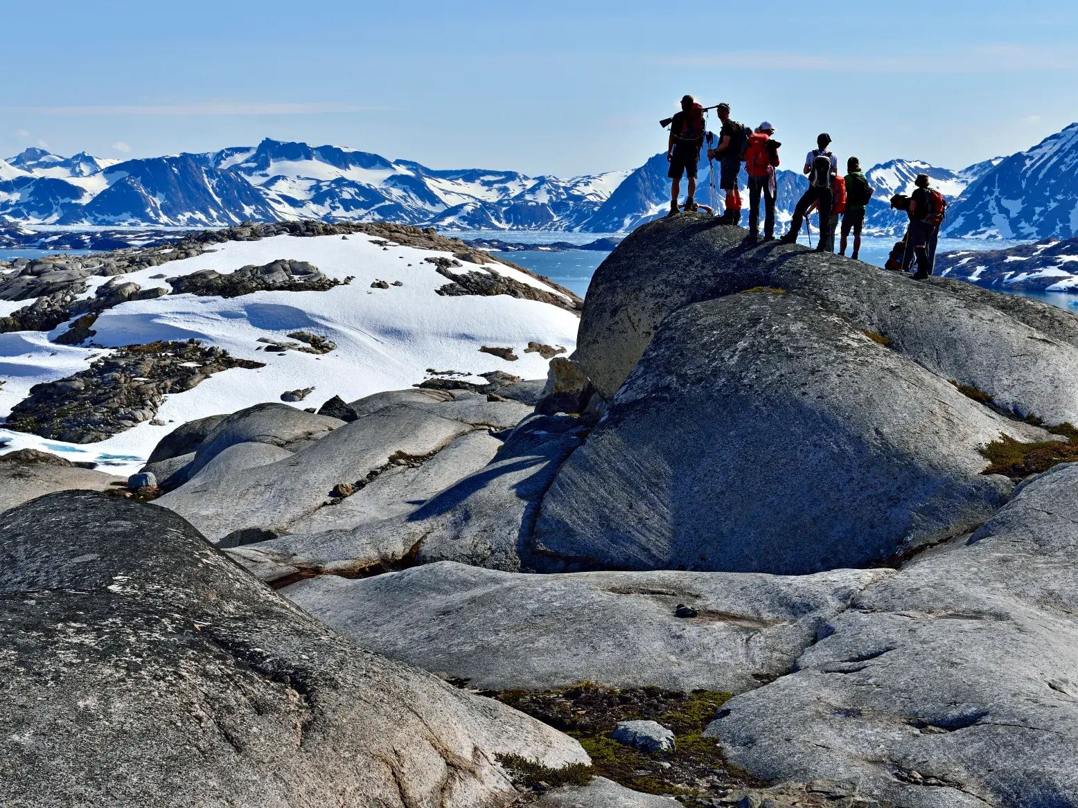 Trekkinggruppe auf einem Felsgipfel in Ostgrönland blickt auf die weite, schneebedeckte Berg- und Fjordlandschaft.