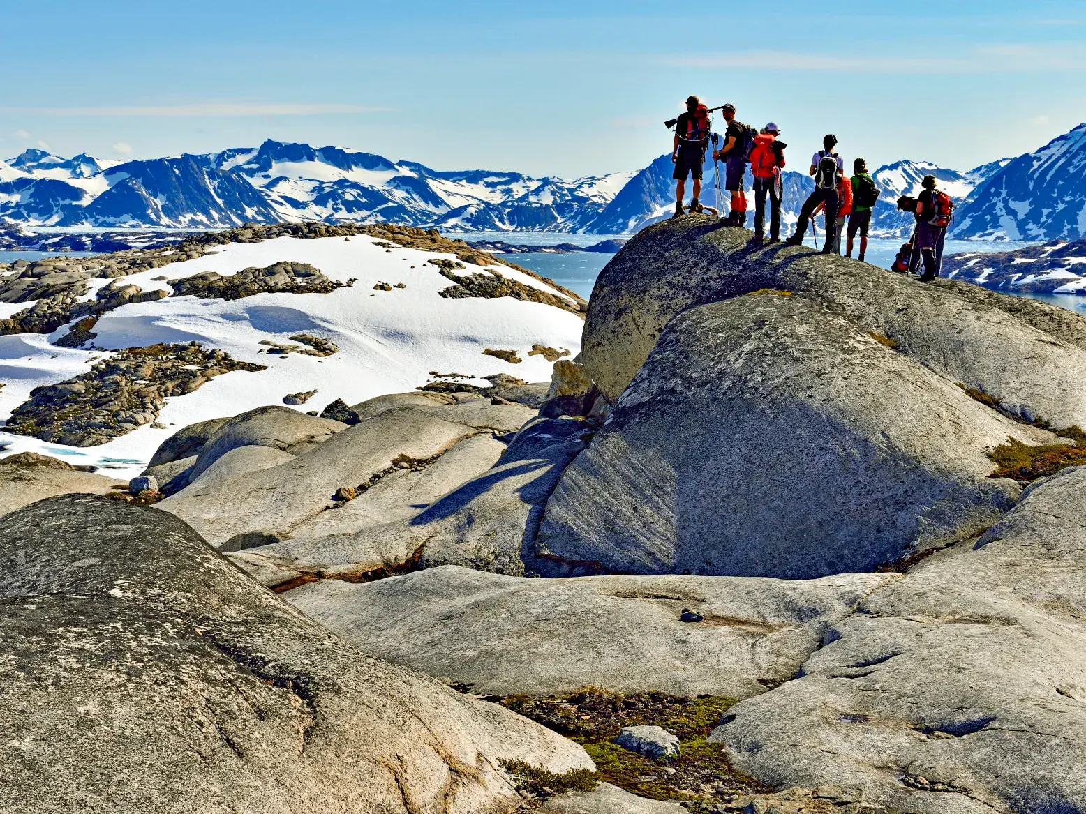 Eine Trekkinggruppe pausiert auf einem Felsen und blickt über die weite Fjordlandschaft der arktischen Wildnis Ostgrönlands.