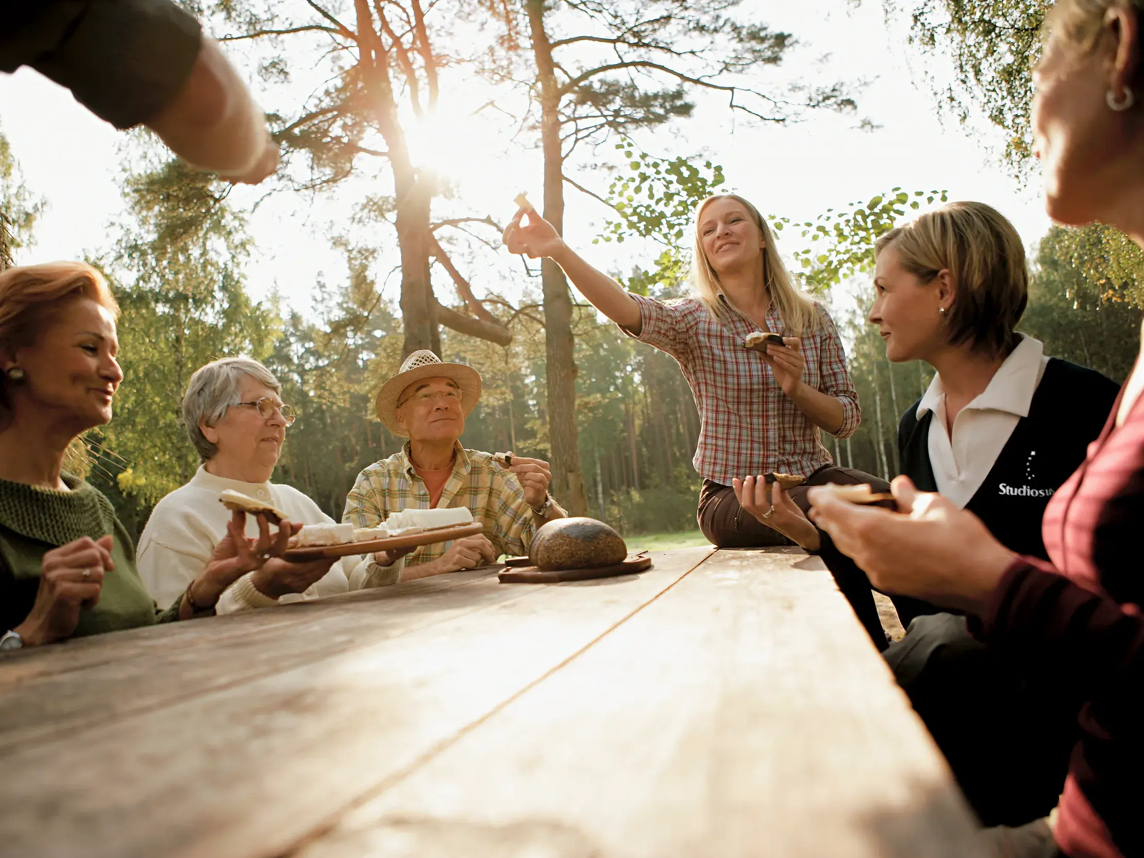 Reisegruppe mit Studiosus Reiseleiterin bei einem sonnigen Picknick an einem Holztisch in einem estnischen Pinienwald.