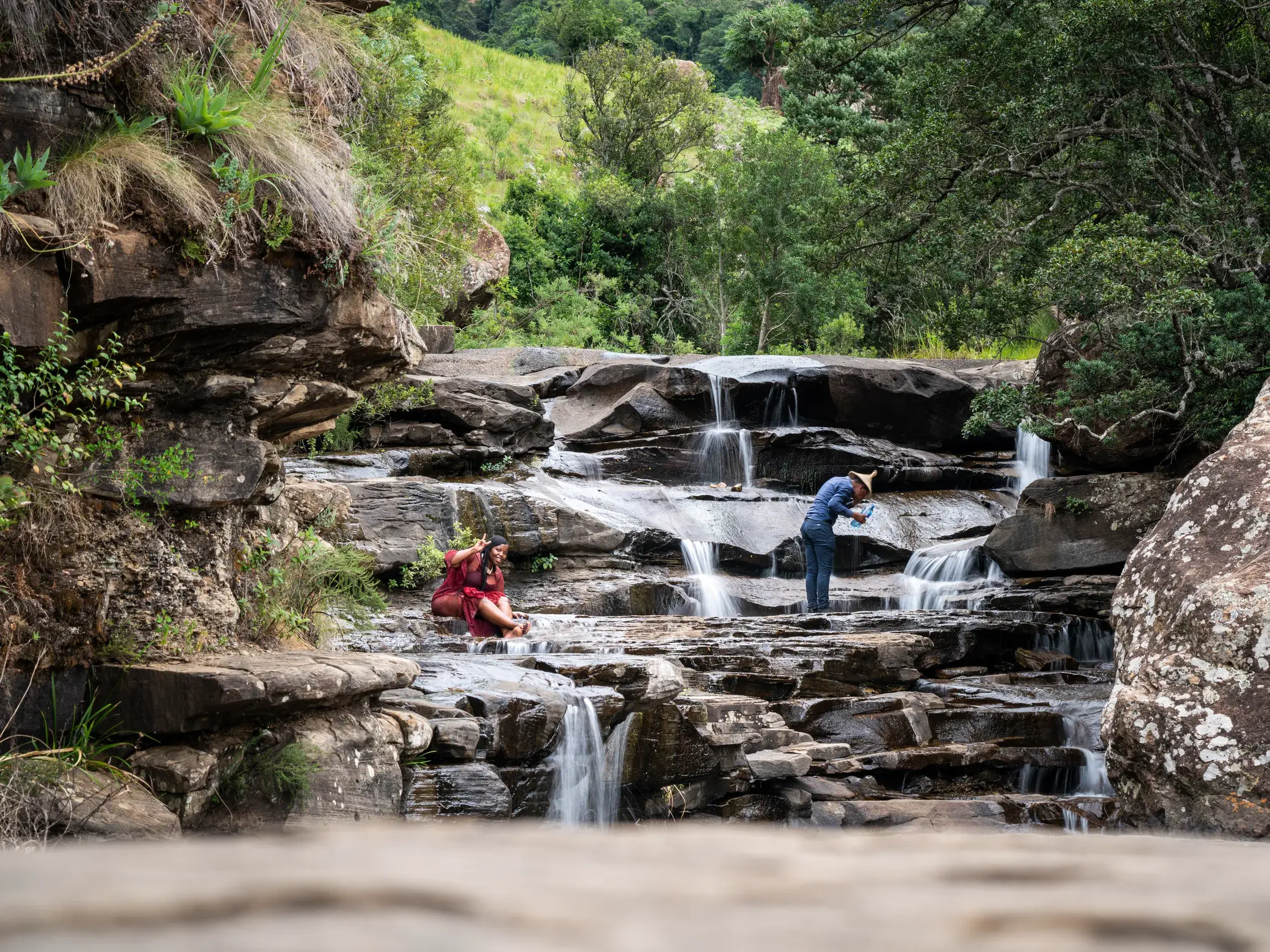 Während einer Wanderpause in Südafrika fotografiert ein Reisender einen stufigen Wasserfall, während eine Frau auf den Felsen entspannt.