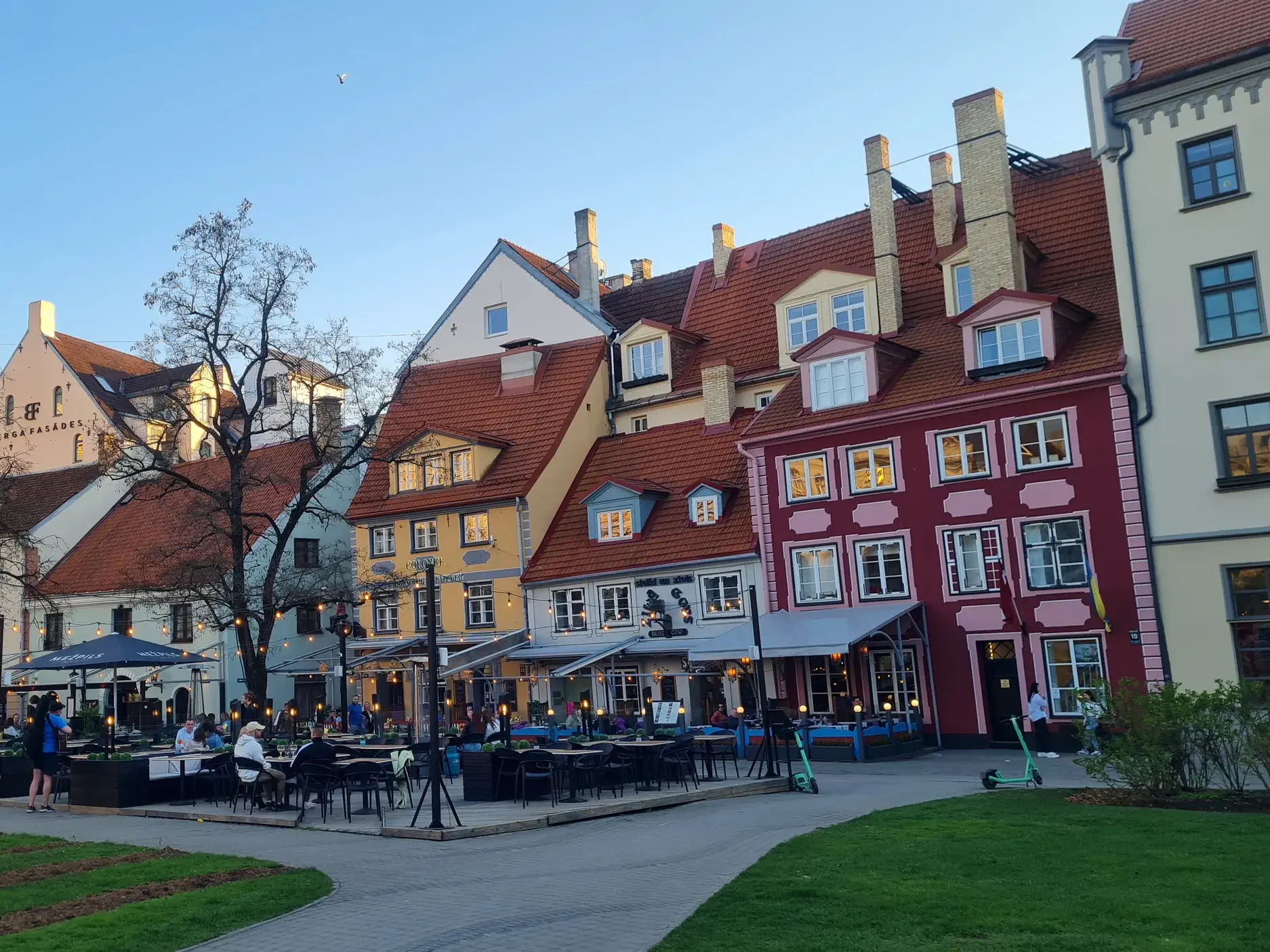 Bunte Giebelhäuser in der Altstadt von Riga mit Straßencafés, im Hintergrund die Turmspitze des Rigaer Doms bei Abendlicht.