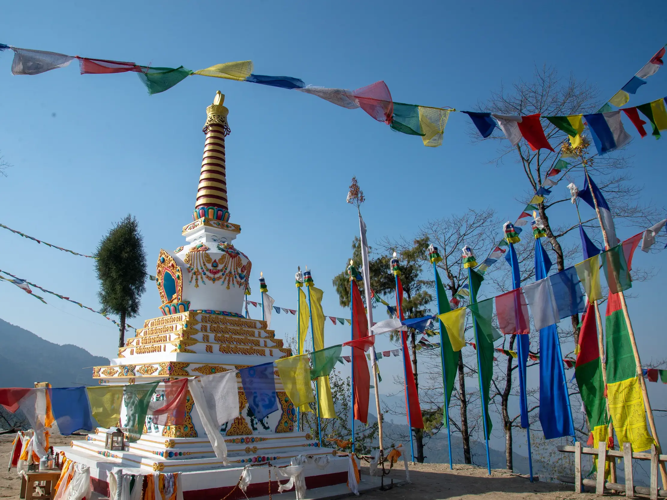 Ein weißer, verzierter Chörten in Bhutan, umgeben von bunten Gebetsfahnen, die vor der Bergkulisse des Himalaya im Wind wehen.