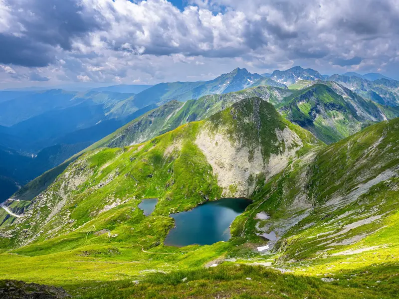 Panoramablick beim Trekking im Fagaras Gebirge: Der blaue Capra-See umgeben von grünen Berghängen und den Karpaten-Gipfeln.