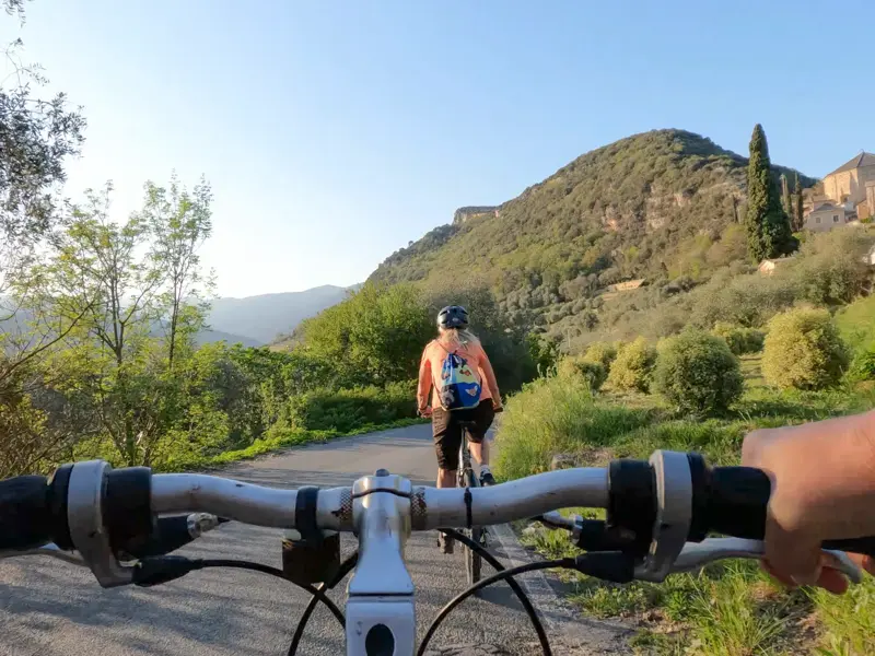 Blick über den Fahrradlenker auf eine Frau, die durch die grüne Hügellandschaft radelt.
