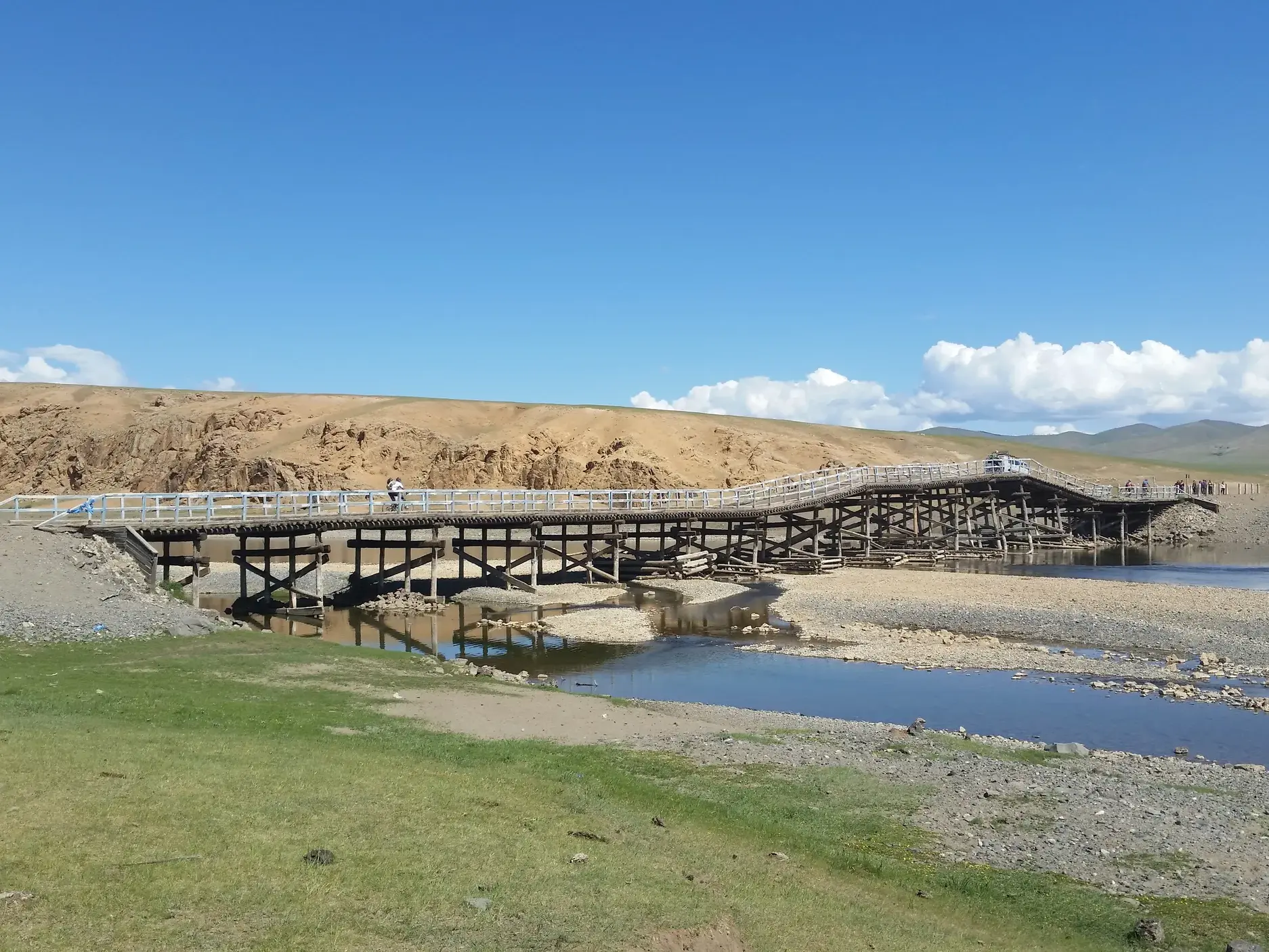 Eine Reisegruppe überquert auf einer Radreise eine lange Holzbrücke über ein Flussbett in der weiten Steppe der Mongolei.