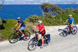 Eine kleine Reisegruppe mit E-Bikes radelt auf einer Küstenstraße auf den Kykladen entlang des tiefblauen Meeres.