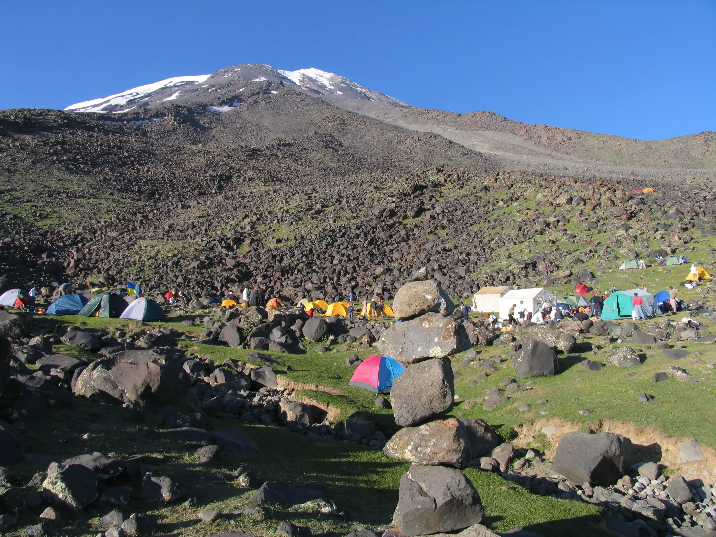 Belebtes Basislager mit bunten Zelten und einer Trekking-Gruppe an den steinigen Hängen des schneebedeckten Ararat.