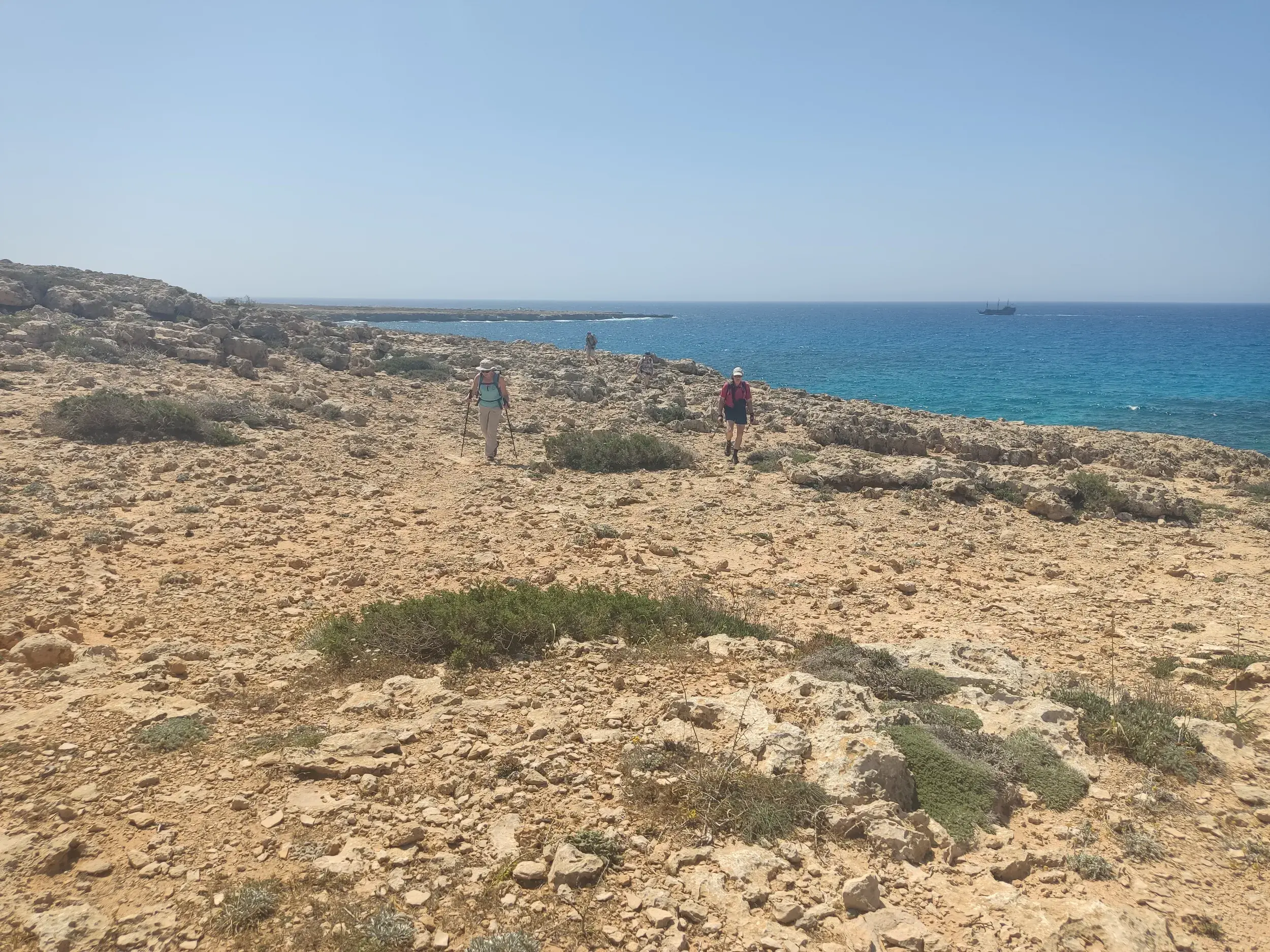 Eine kleine Wandergruppe wandert mit Stöcken auf einem felsigen Pfad entlang der Küste am Kap Greco auf Zypern bei Sonnenschein.