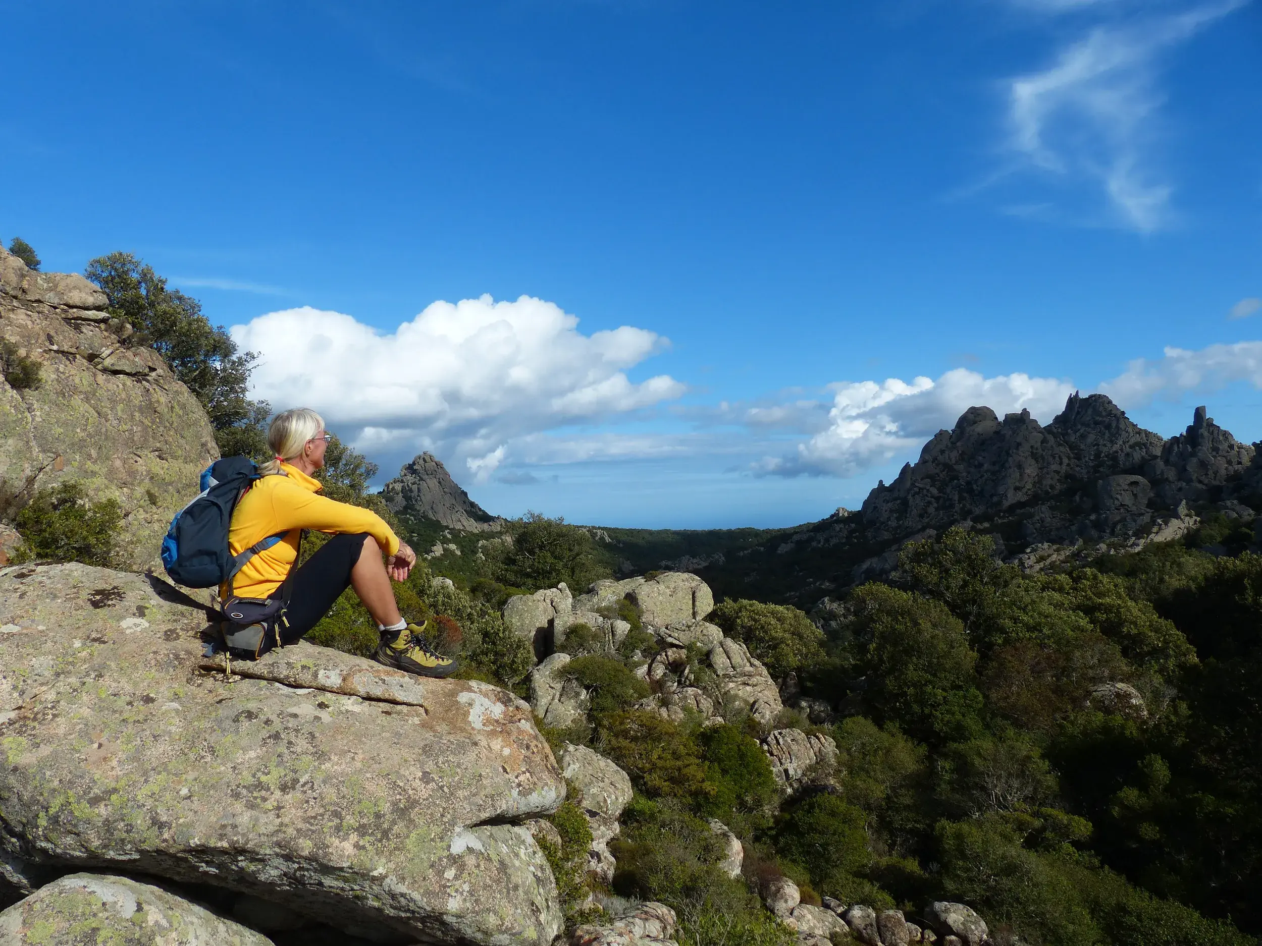 Wanderin in gelber Jacke ruht auf einem Granitfelsen und blickt über die Berglandschaft des Naturschutzgebiets I Sette Fratelli.