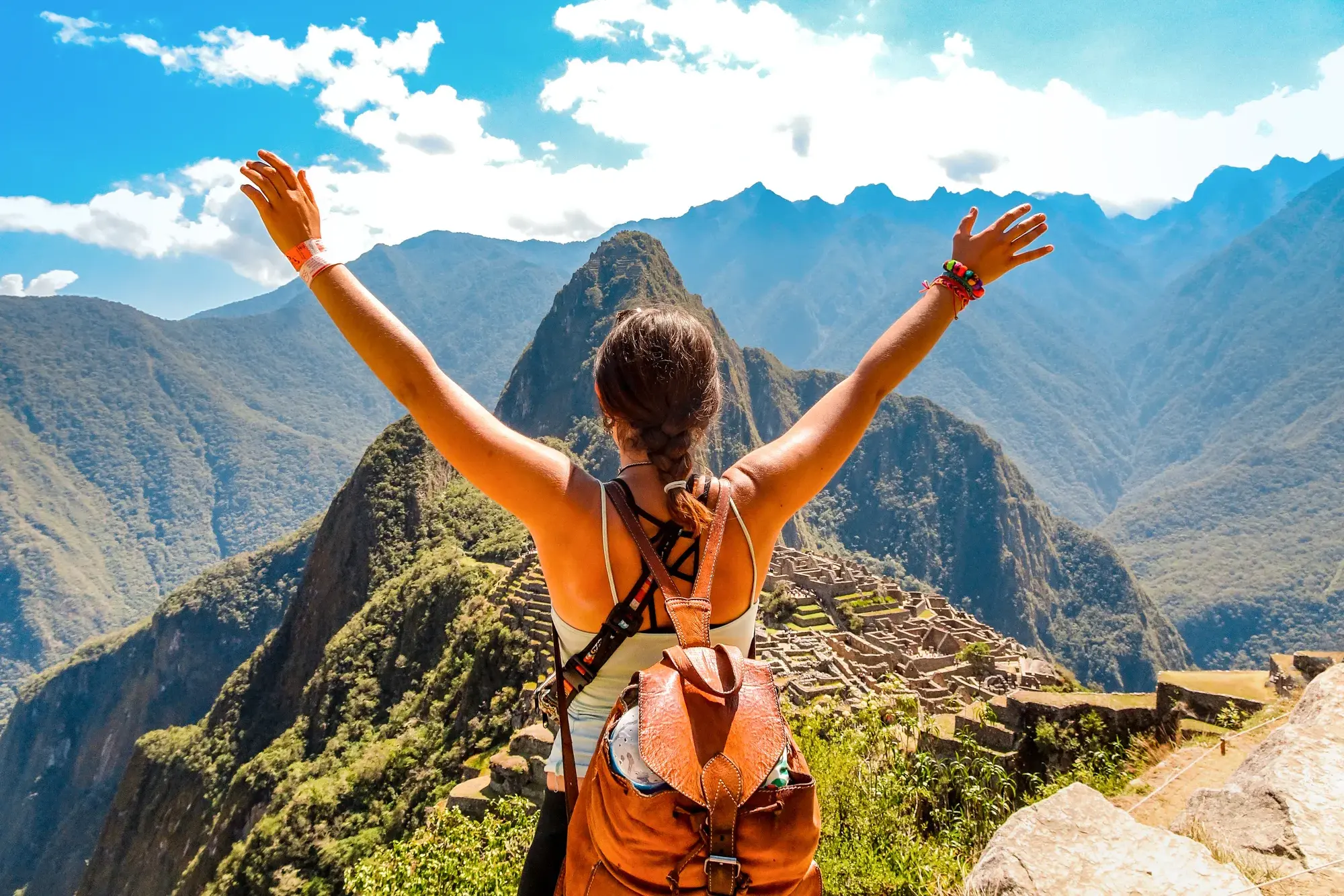 Junge Frau mit Rucksack streckt jubelnd die Arme hoch und blickt auf die beeindruckende Inkastadt Machu Picchu in den Anden.