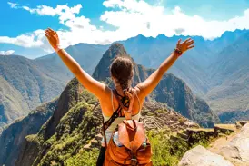 Junge Frau mit Rucksack streckt jubelnd die Arme hoch und blickt auf die beeindruckende Inkastadt Machu Picchu in den Anden.