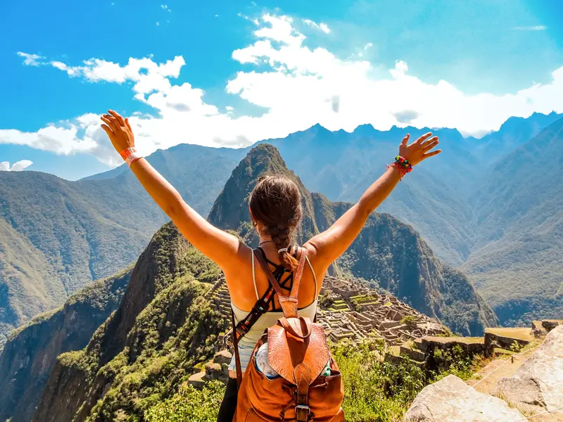 Junge Frau mit Rucksack streckt jubelnd die Arme hoch und blickt auf die beeindruckende Inkastadt Machu Picchu in den Anden.