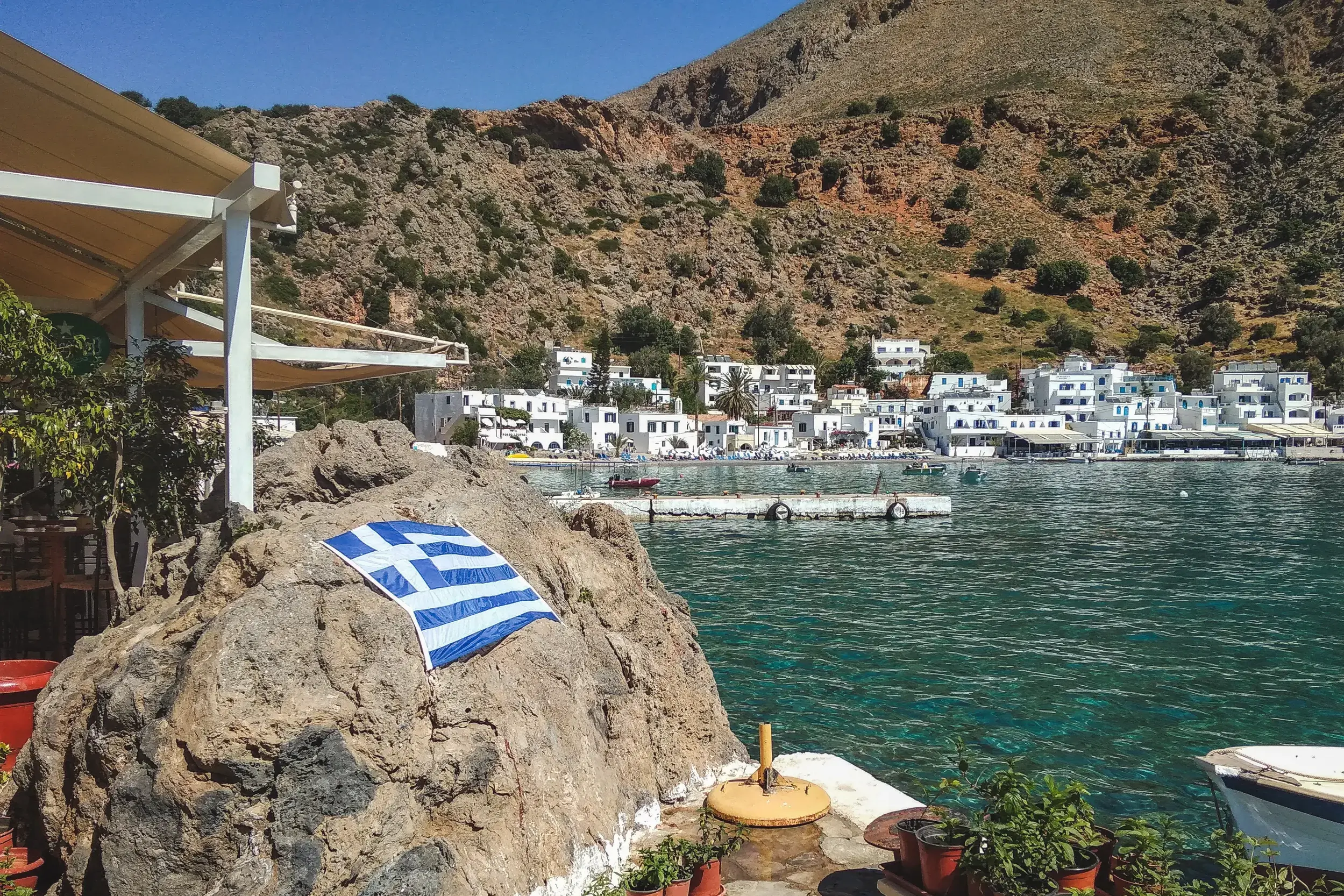 Das weiße Fischerdorf Loutro in seiner Bucht an Kretas Südküste, mit einer griechischen Flagge auf einem Felsen im Vordergrund.