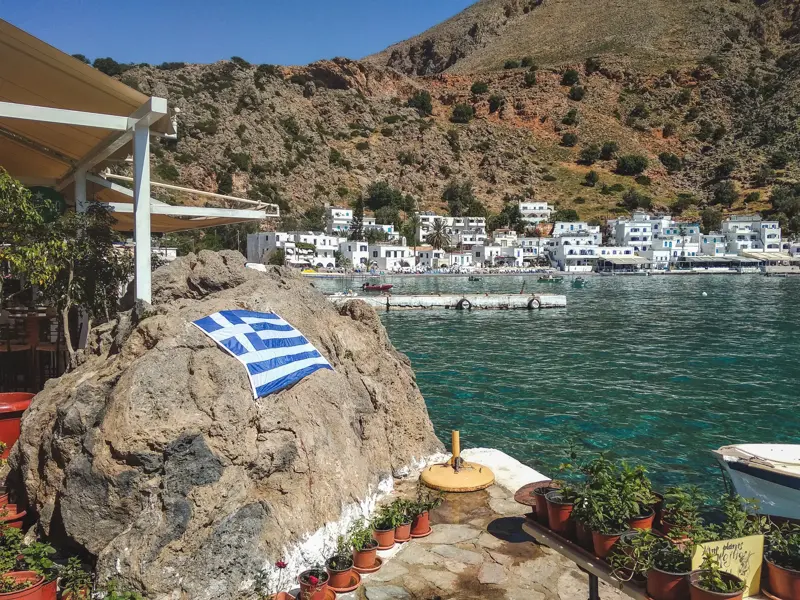 Das weiße Fischerdorf Loutro in seiner Bucht an Kretas Südküste, mit einer griechischen Flagge auf einem Felsen im Vordergrund.
