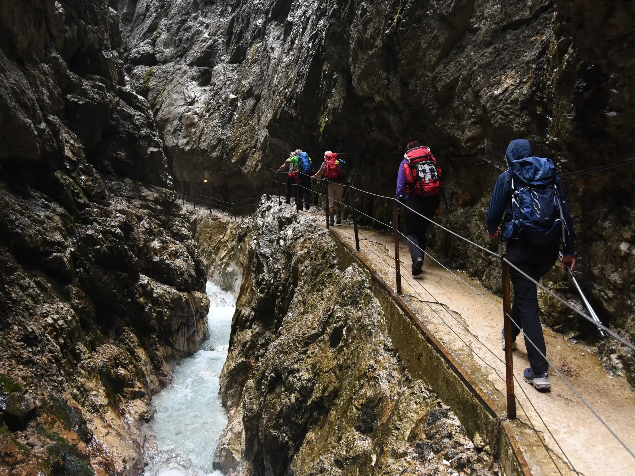 Eine Wandergruppe durchquert auf einem gesicherten Steg die enge Höllentalklamm neben einem reißenden Wildbach.