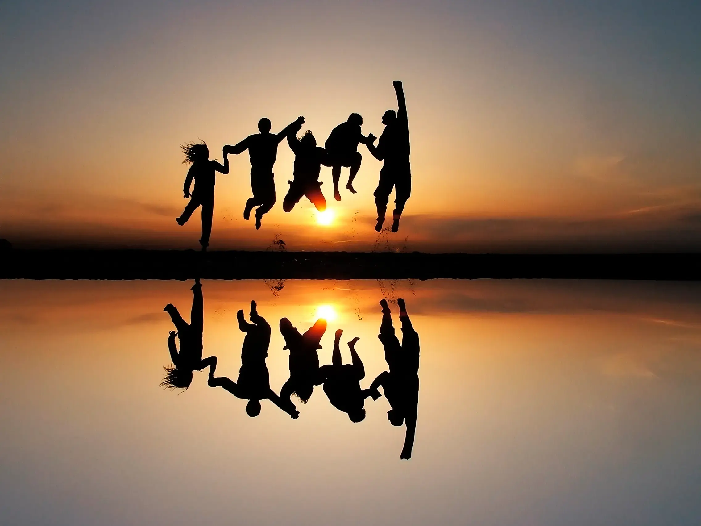 Silhouetten einer jungen Reisegruppe, die bei Sonnenuntergang am Salar de Atacama in die Luft springt und sich im Wasser spiegelt.