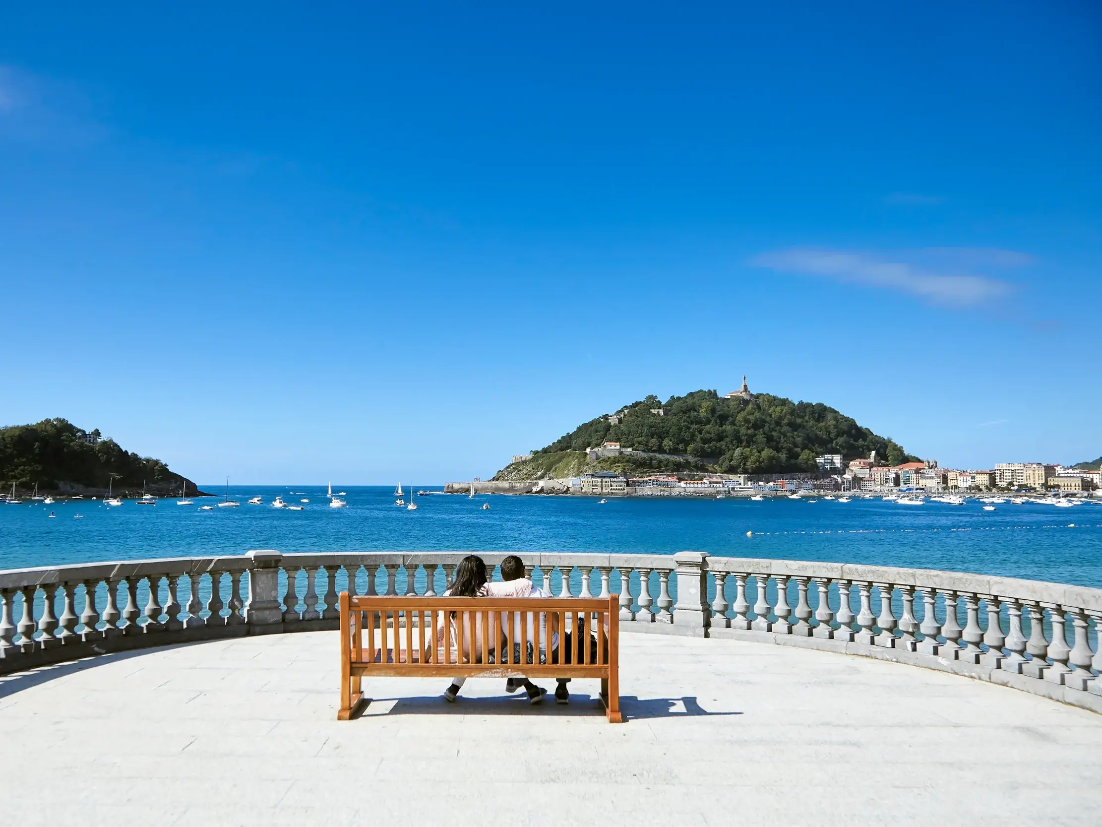 Zwei Personen auf einer Bank genießen den Blick auf die muschelförmige Bucht La Concha in San Sebastián mit dem Monte Urgull.