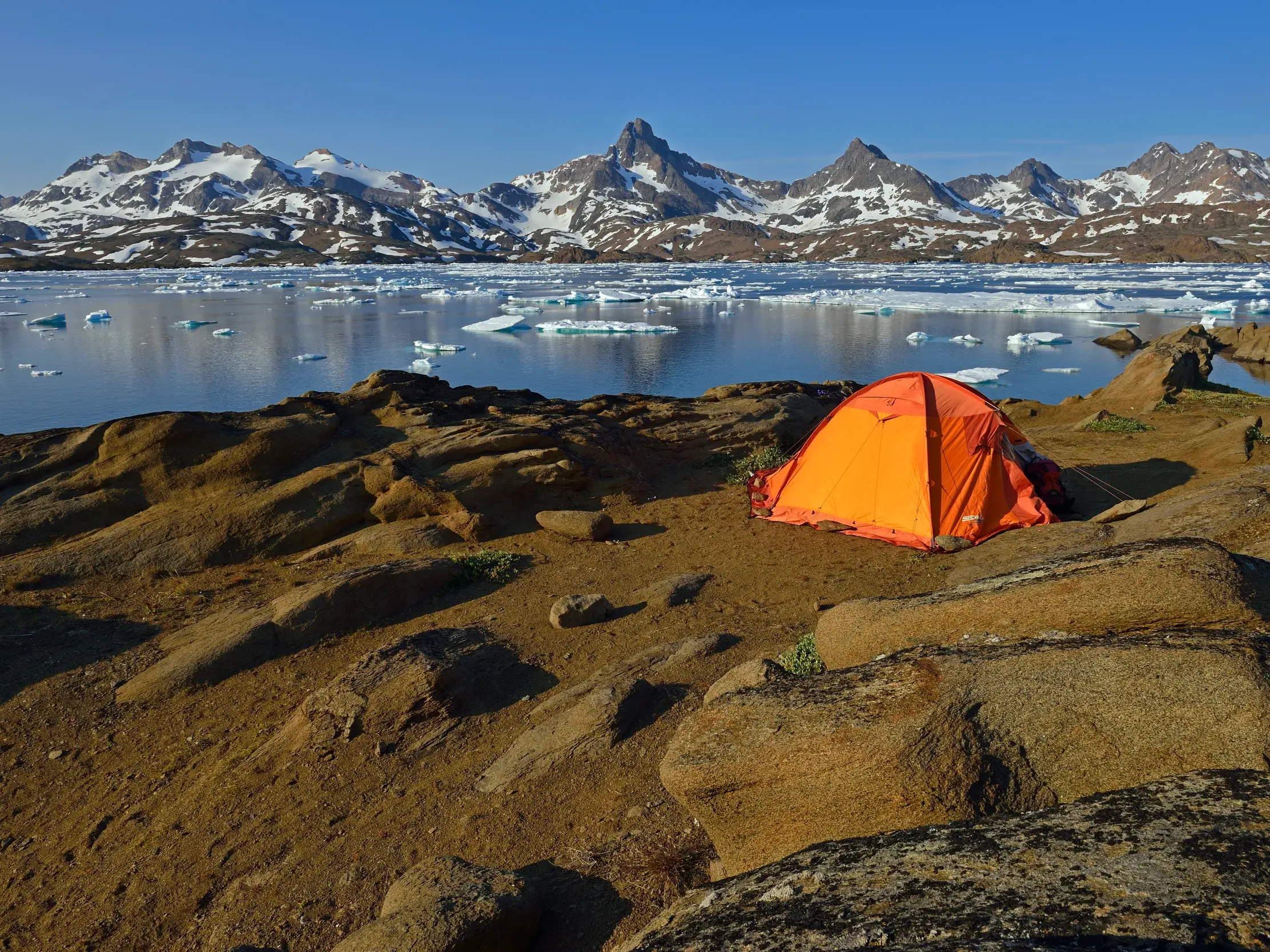 Ein orangefarbenes Zelt steht beim Trekking auf felsigem Grund am Ufer des Johan-Petersen-Fjords mit Blick auf Eisberge in Grönland.