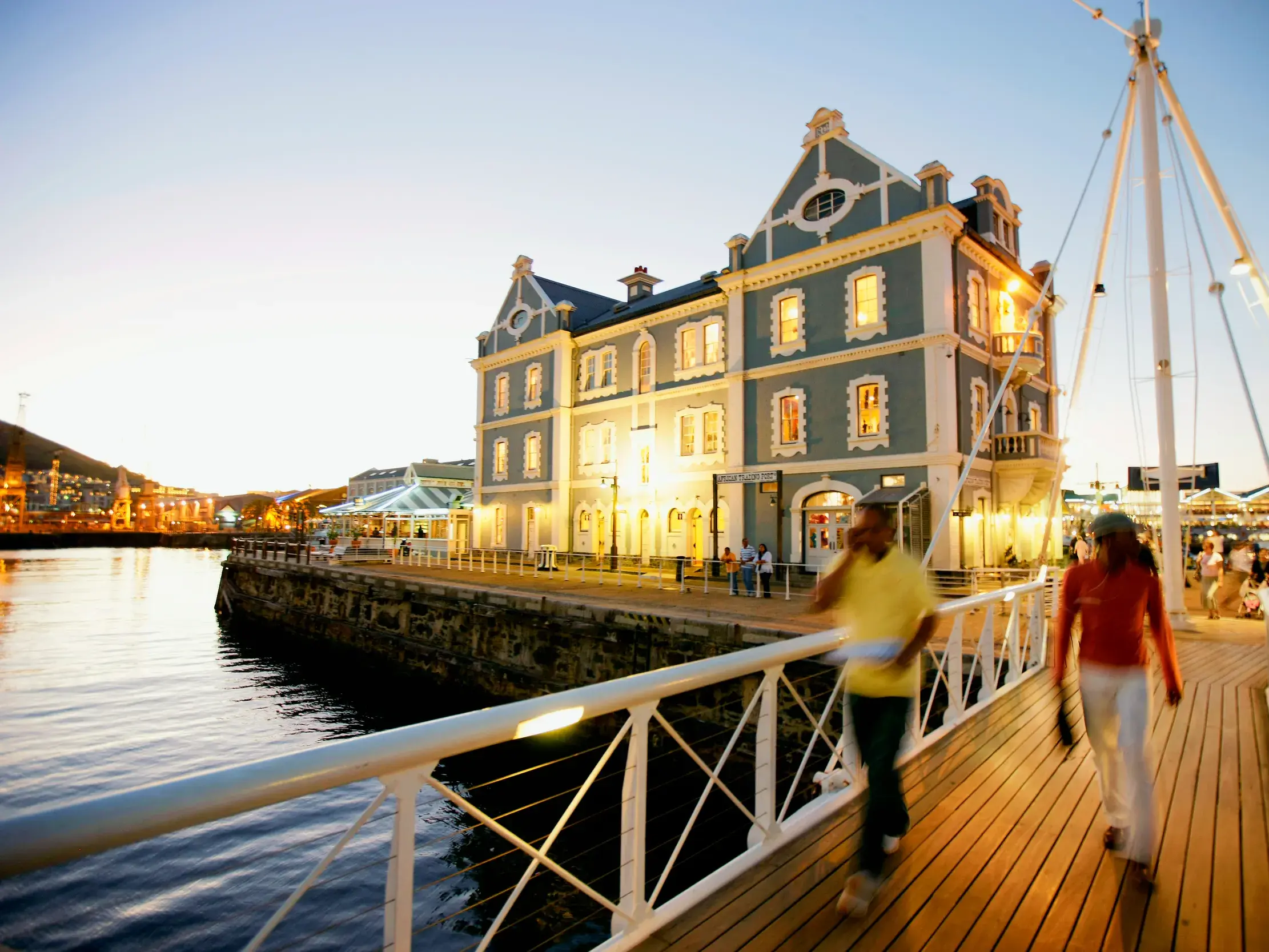 Ein Paar genießt den Abend auf einer Rundreise durch Südafrika an der belebten V&A Waterfront in Kapstadt vor historischer Architektur.
