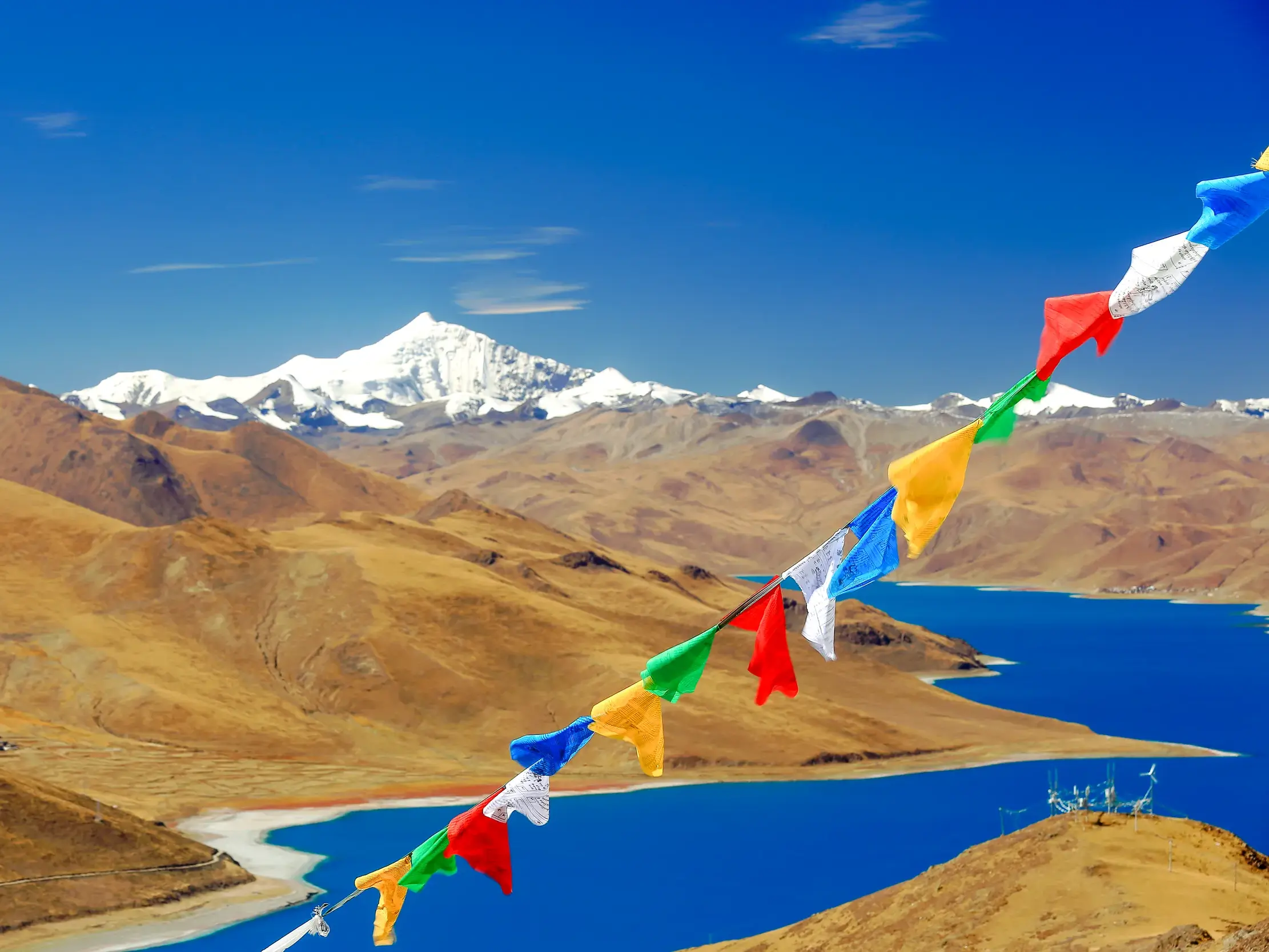 Bunte Gebetsfahnen wehen am Pass Kampa-La mit Blick auf den türkisblauen See Yamdrok-Tso und schneebedeckte Berge in Tibet.