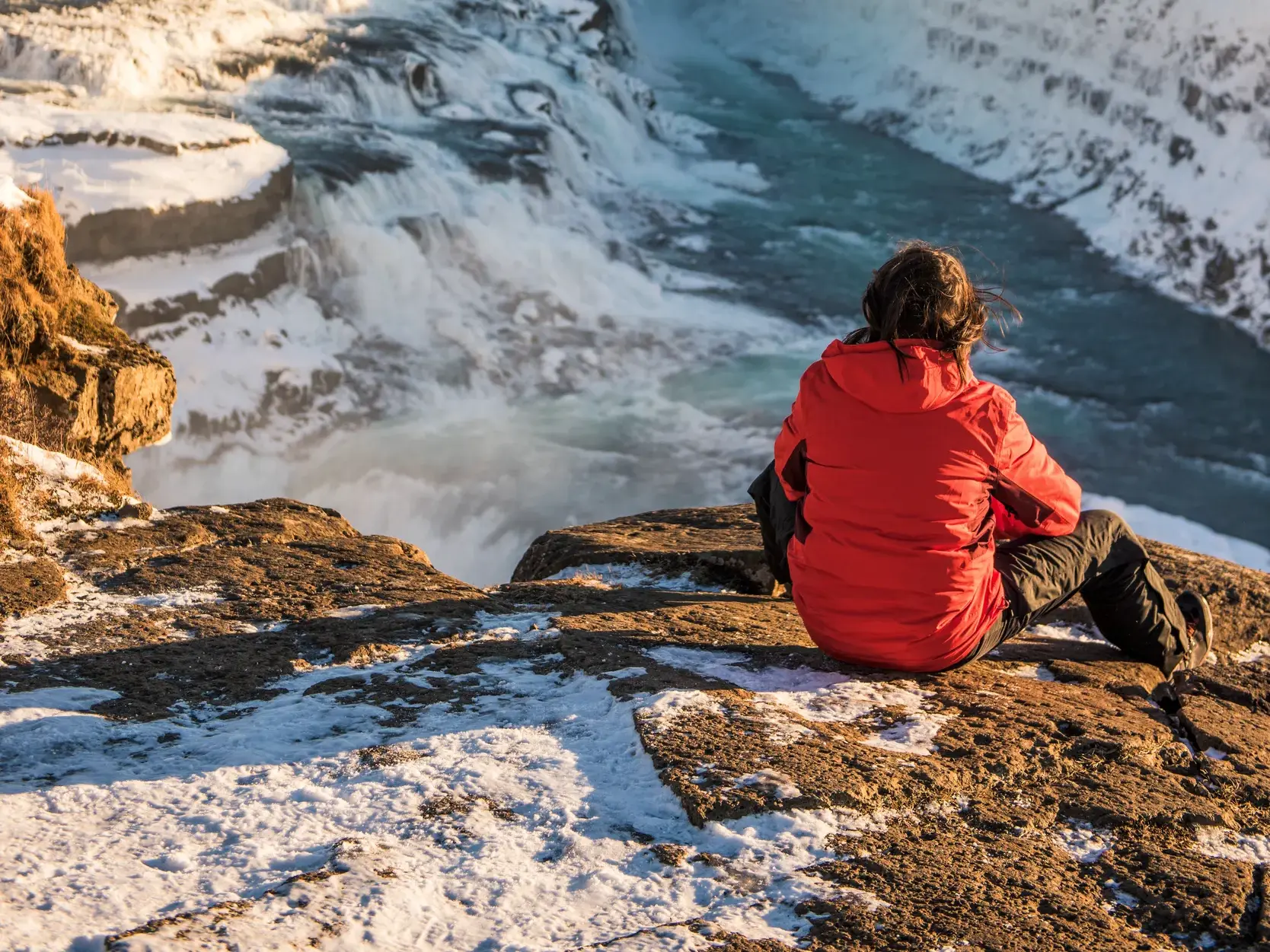 Reisende blickt im Winter von einem verschneiten Felsen auf die tosenden Kaskaden des Wasserfalls Gullfoss in Island.