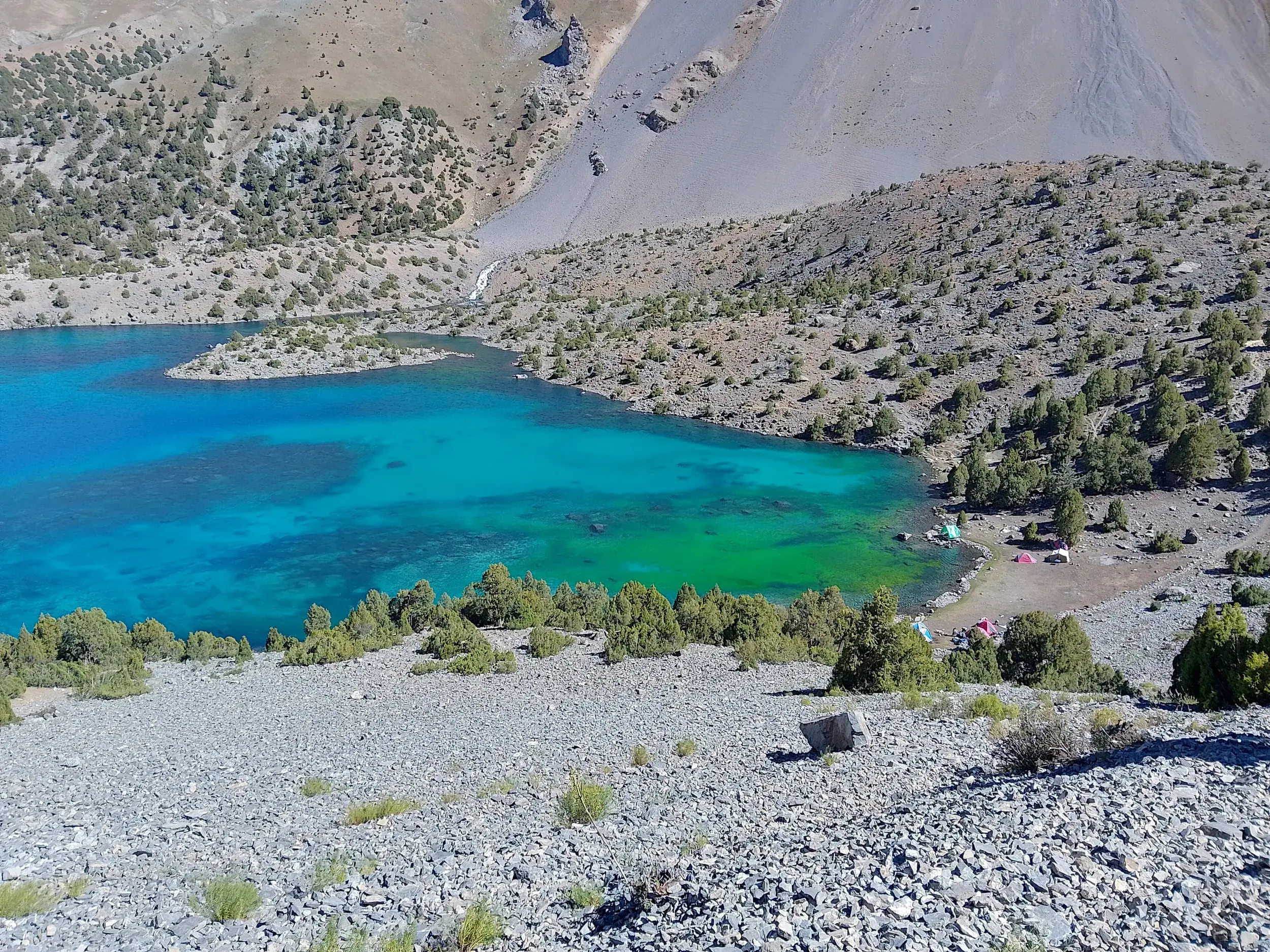 Blick von einem Schotterhang auf den türkisfarbenen Alaudin-See, an dessen Ufer das Zeltlager einer Trekkinggruppe steht.