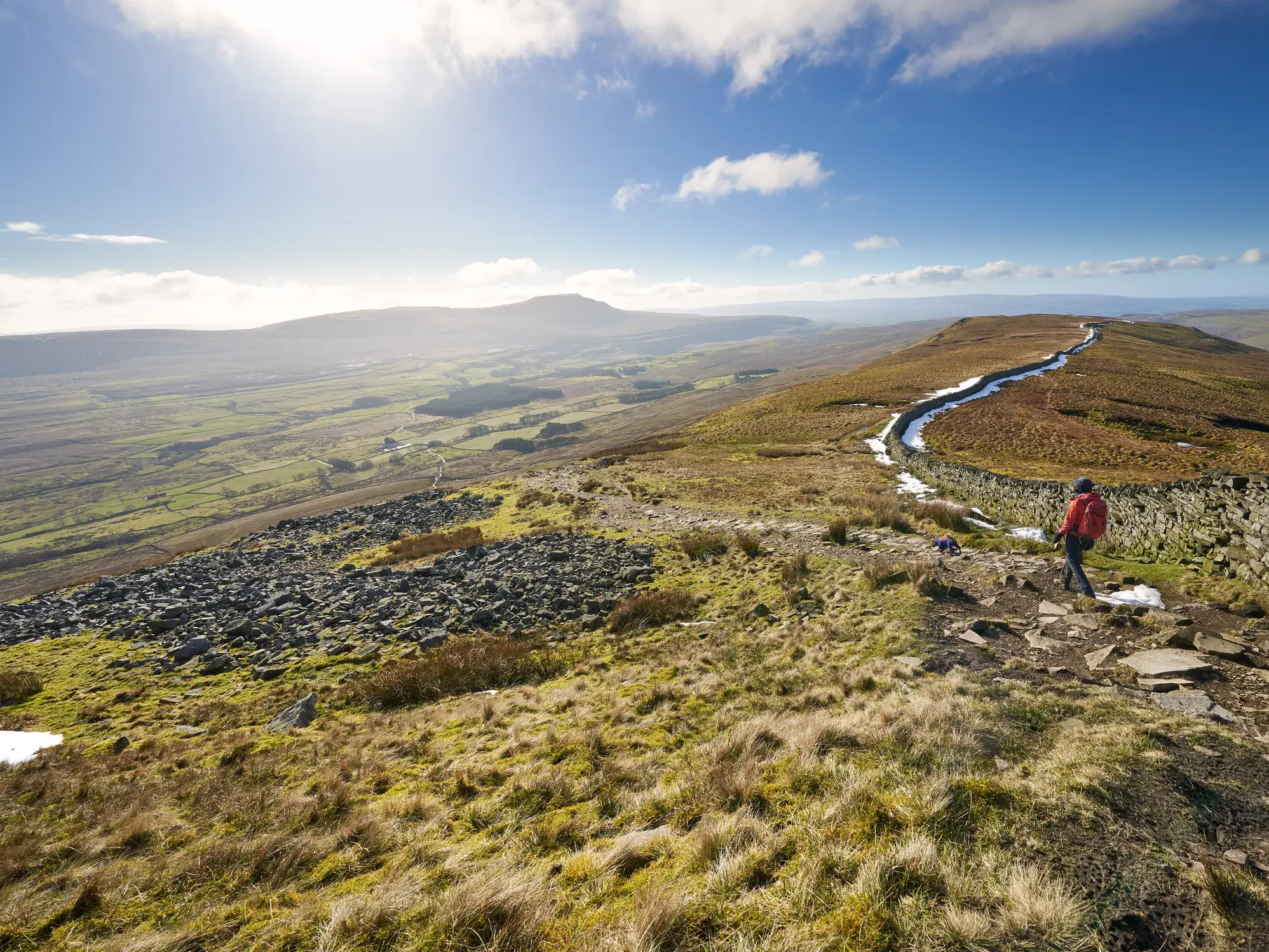 Ein Wanderer auf dem Coast to Coast Walk in England, entlang einer Steinmauer mit Blick über die grünen Hügel des Yorkshire Dales.