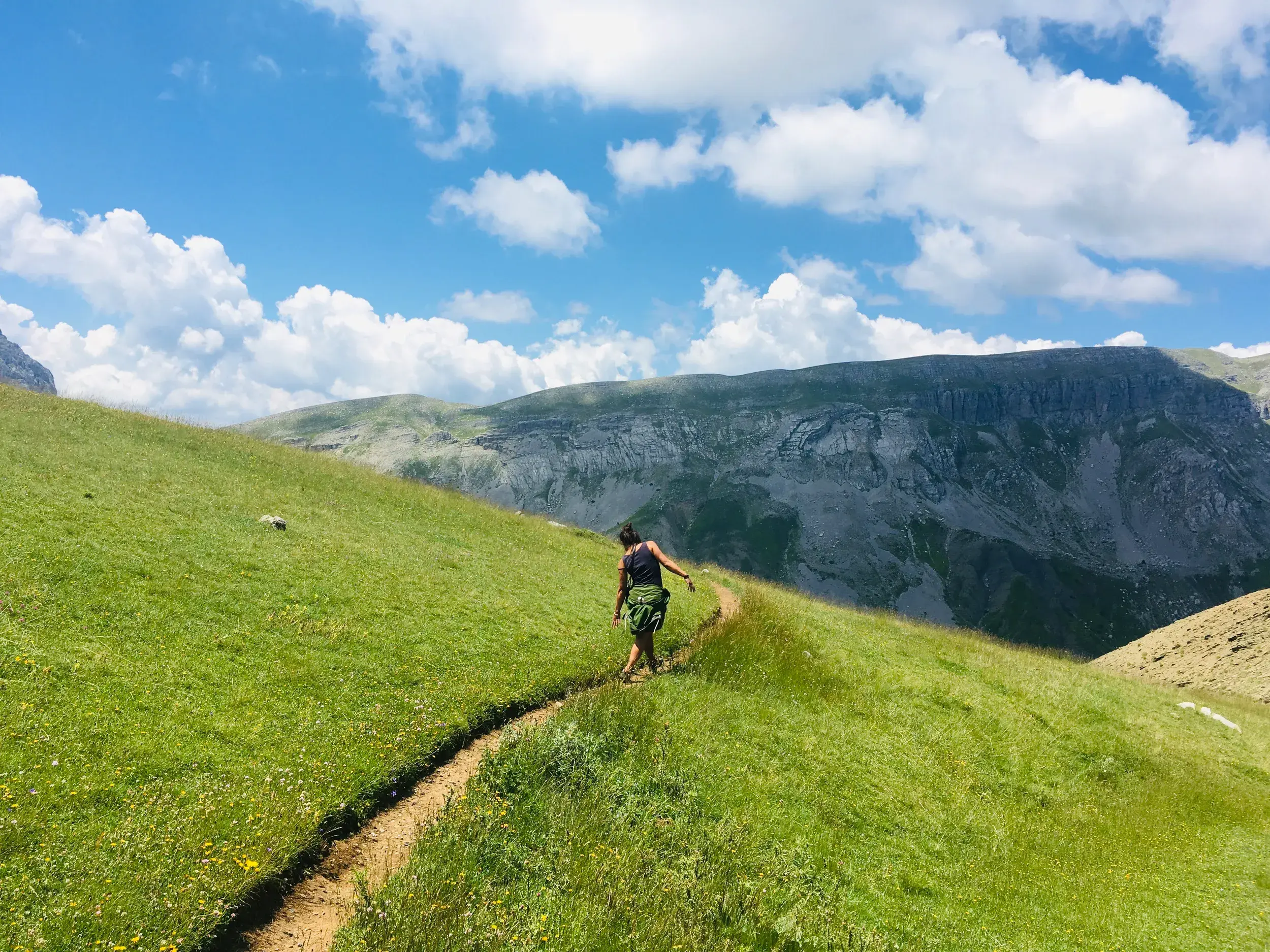 Eine Wanderin folgt einem schmalen Pfad über eine saftig grüne Almwiese im Pindos-Gebirge während einer Trekkingreise in der Zagori-Region.