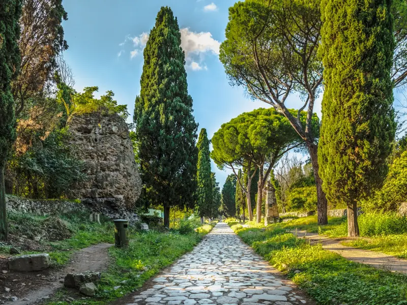 Wanderweg auf dem antiken Pflaster der Via Appia Antica, vorbei an Ruinen, gesäumt von Zypressen und Pinien bei Rom.