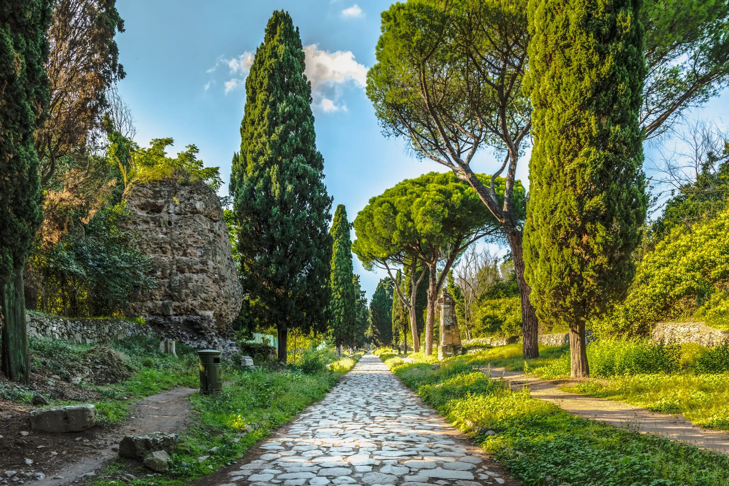 Wanderweg auf der alten Römerstraße Via Appia Antica bei Rom, gesäumt von hohen Zypressen, Pinien und antiken Ruinen.